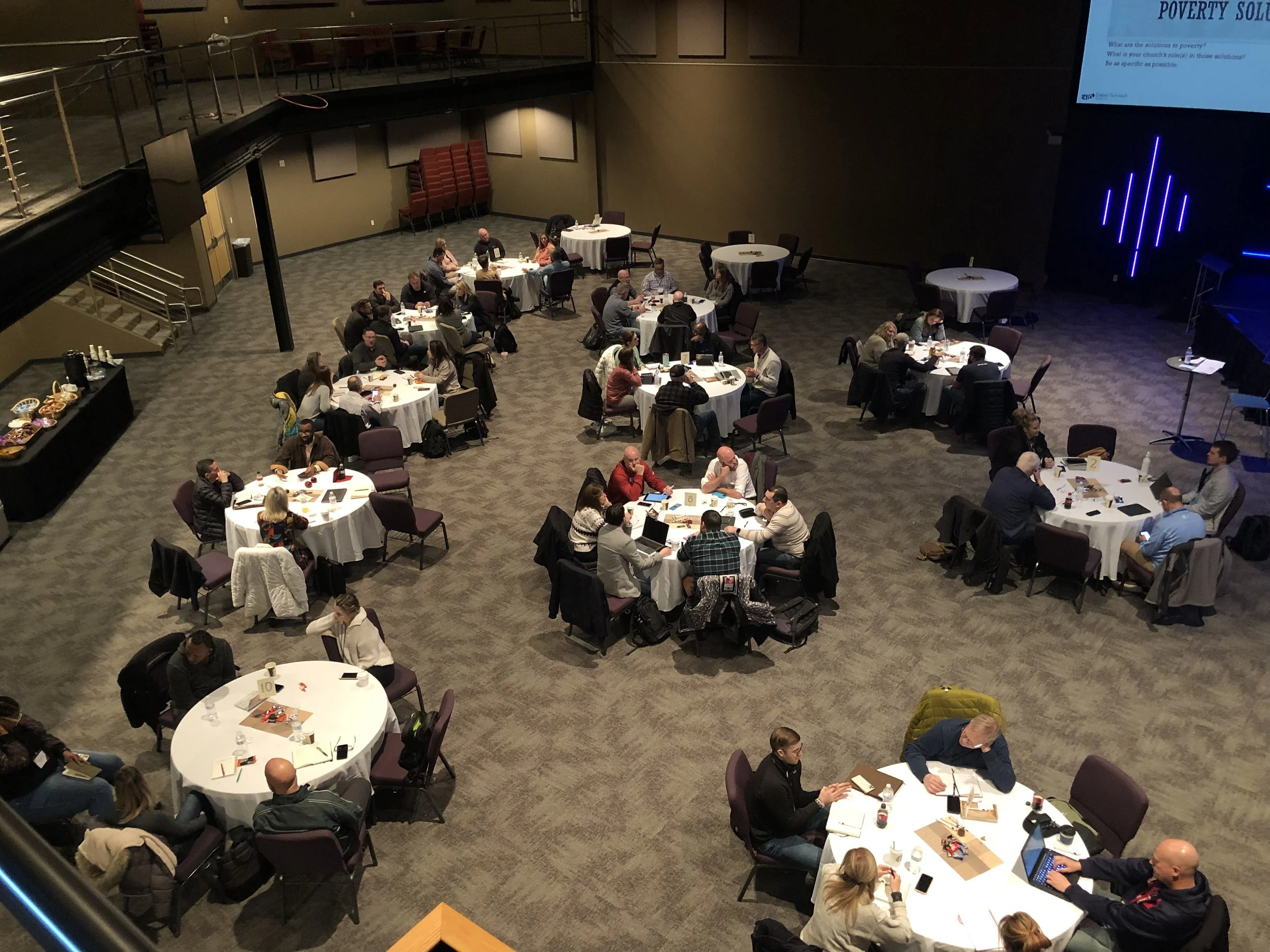 Conference or workshop attendees seated at round tables in a large room, some working on laptops, with food and beverages on the tables, and a presentation screen on the wall.