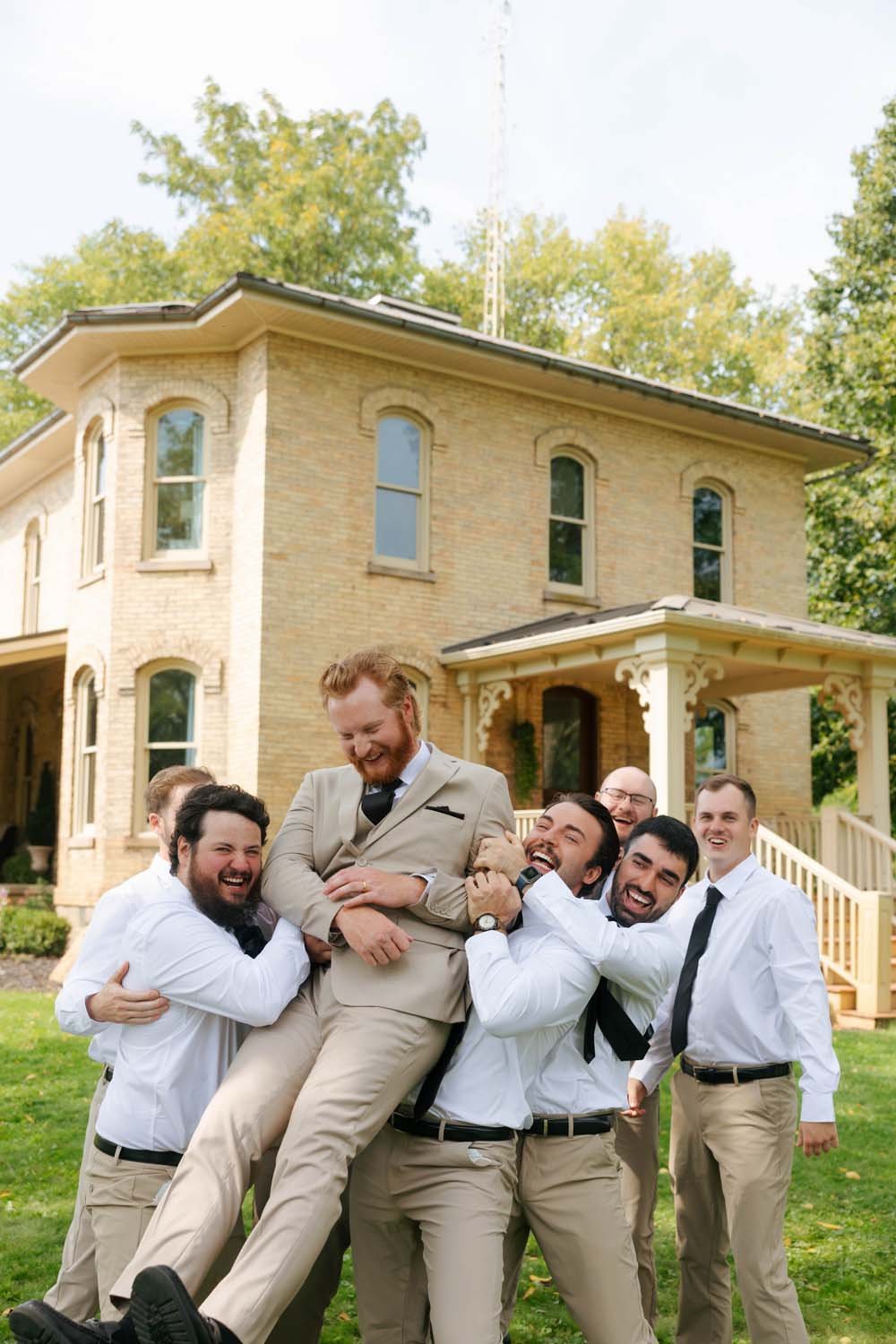 A group of men dressed in beige dress pants and white shirts lifting a man in a beige suit in the air in front of a large brick house with trees in the background.