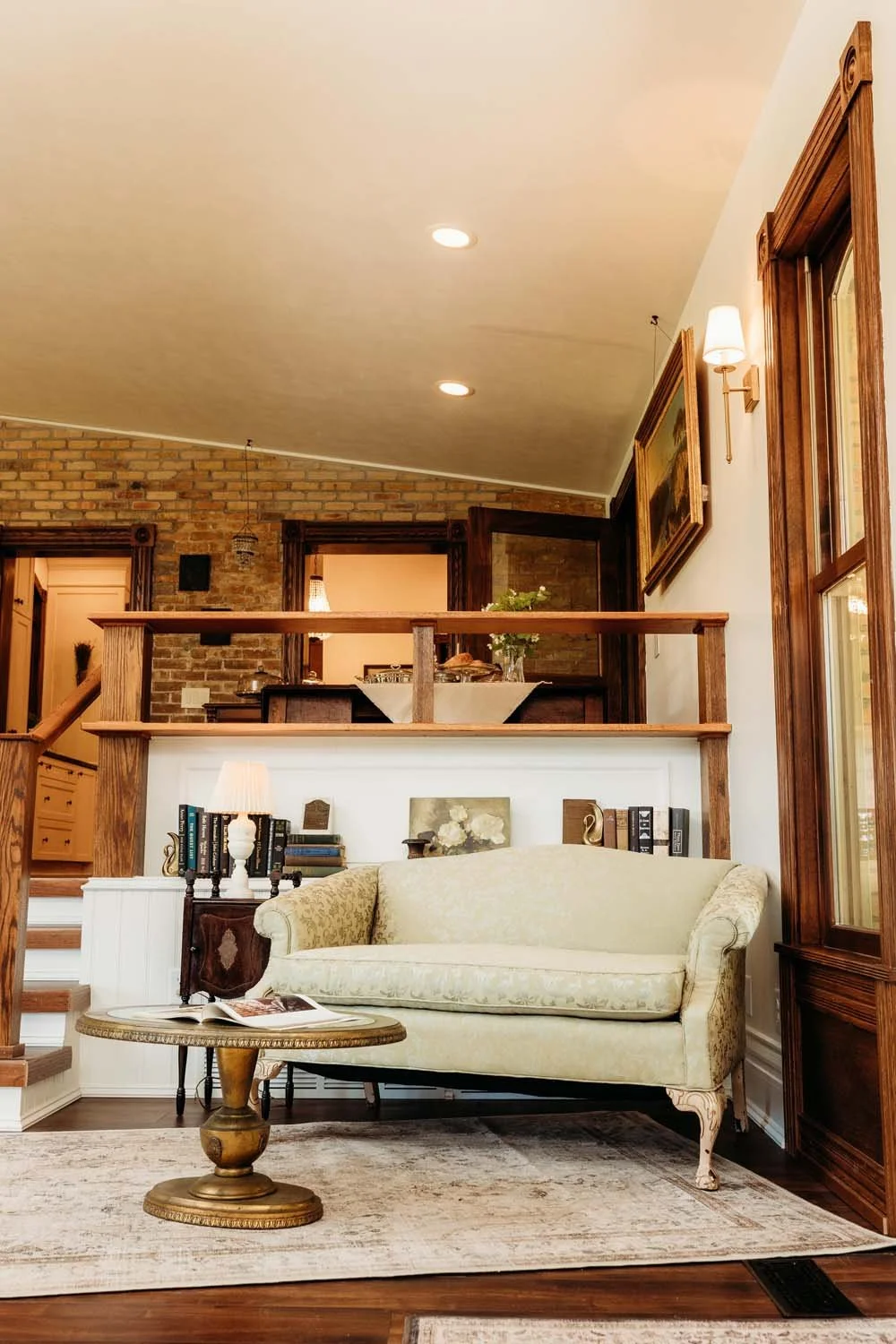 Living room with vintage cream sofa, wooden side table with books and lamp, brick wall, wooden railing, and open kitchen in background.