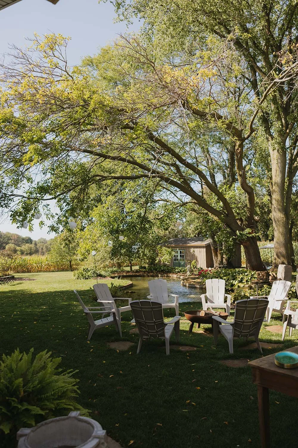A backyard scene with white Adirondack chairs arranged around a fire pit, a pond with a small waterfall, large trees providing shade, and a wooden shed in the background under a partly cloudy sky.