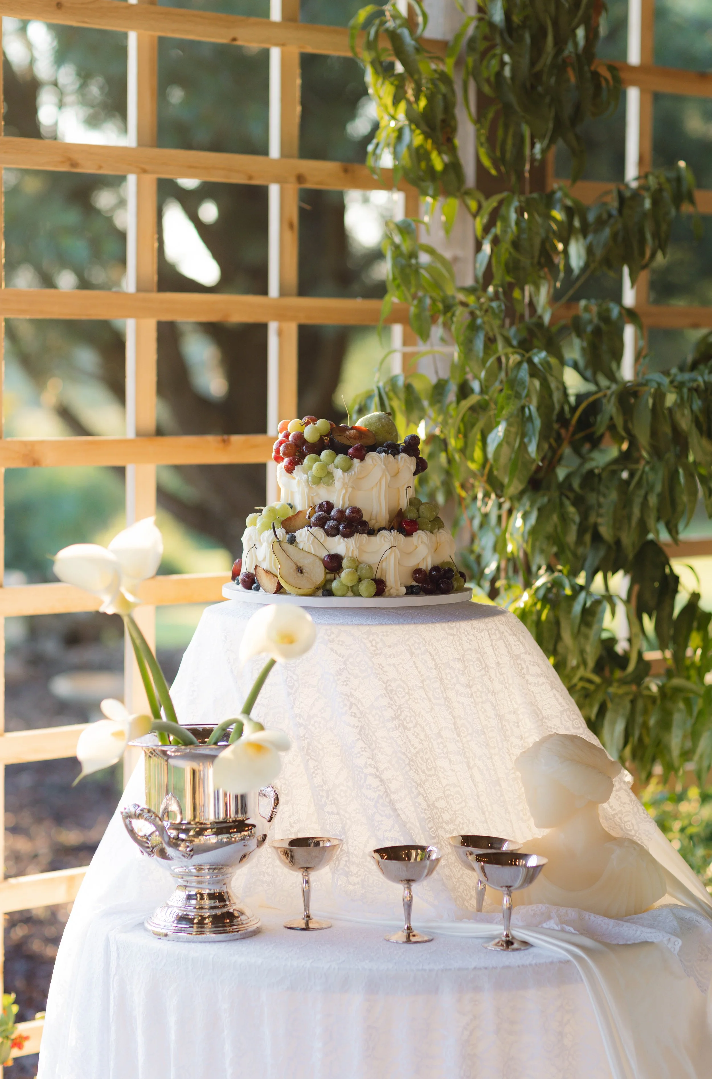 A decorated table with a multi-tiered cake topped with various fruits, a silver vase with white calla lilies, and four silver bowls, set against a wooden trellis and lush greenery outdoors.