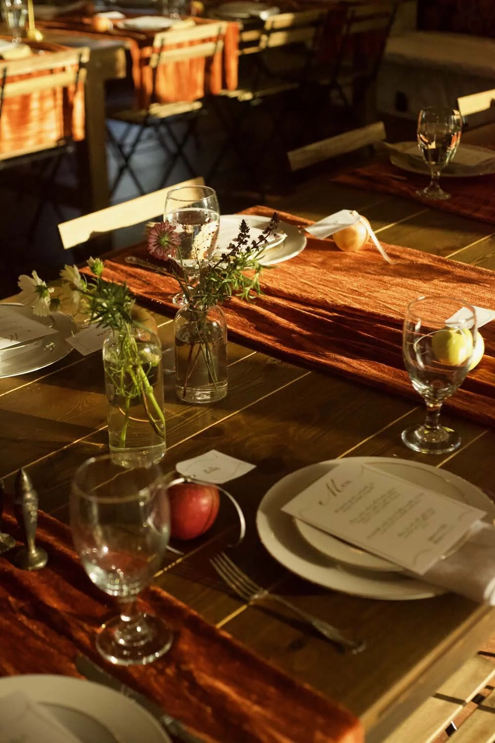 A rustic dining table set for a meal with plates, glasses, cutlery, small flower arrangements, and a menu, with warm lighting and chairs in the background.