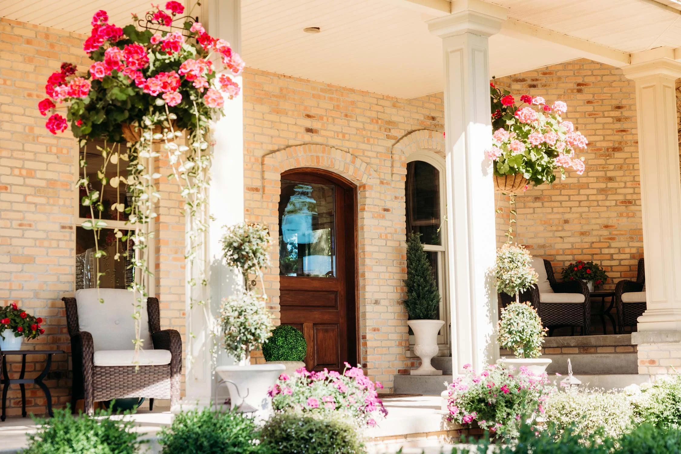 A porch with hanging flower baskets, potted plants, and outdoor furniture in front of a brick house.