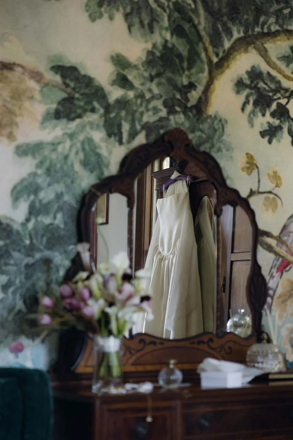 A vintage dresser with a decorative mirror reflecting a cream-colored dress hanging on a wooden hanger, with a floral wallpaper background, a vase of flowers, and small decorative bottles on the dresser.
