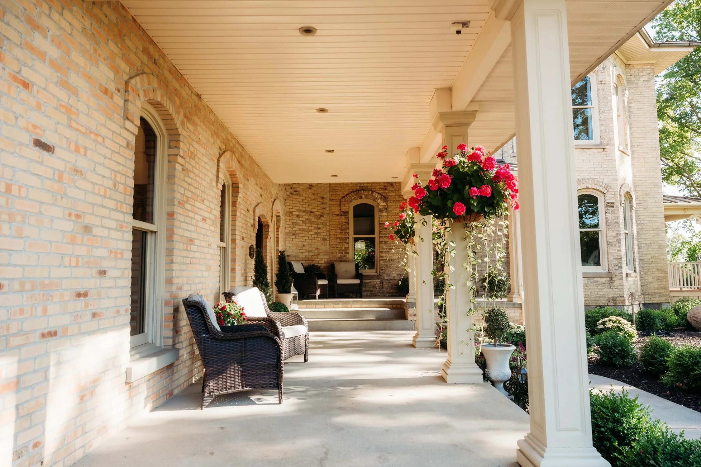 Front porch of a brick house with outdoor seating, potted plants, and hanging flower baskets, overlooking a garden.