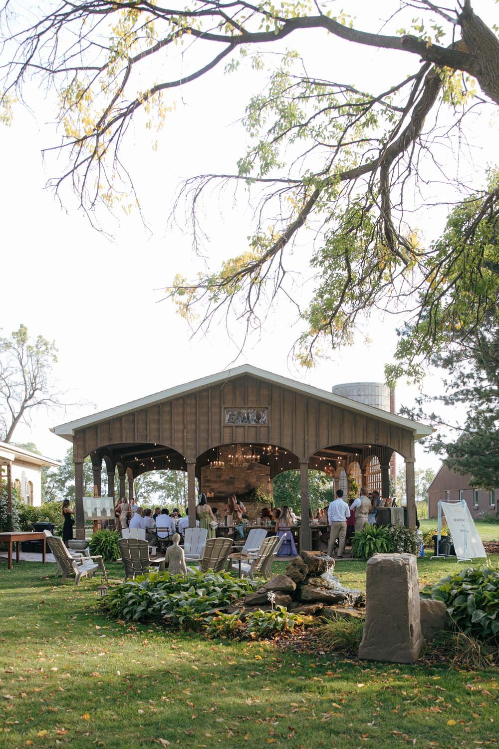 An outdoor wedding reception with guests seated under a wooden pavilion, surrounded by chairs and greenery, with trees and a firepit in the foreground.