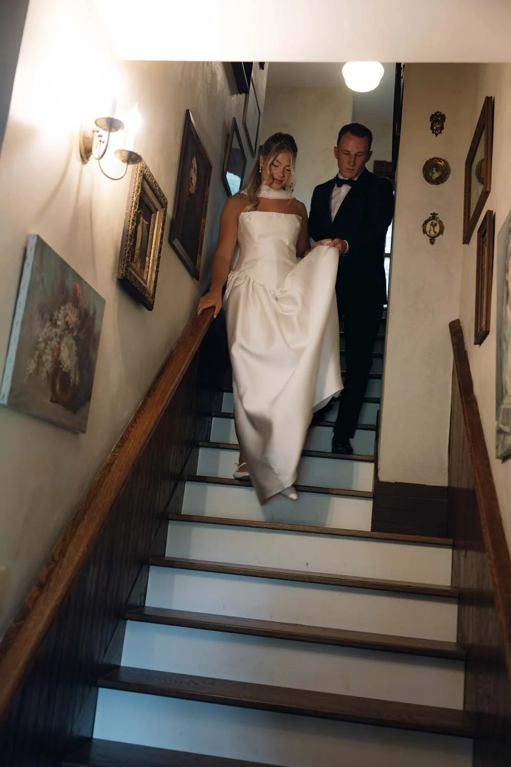 A bride in a white wedding dress being guided down a staircase by a groom in a tuxedo, inside a vintage-style home decorated with framed artwork and wall ornaments.