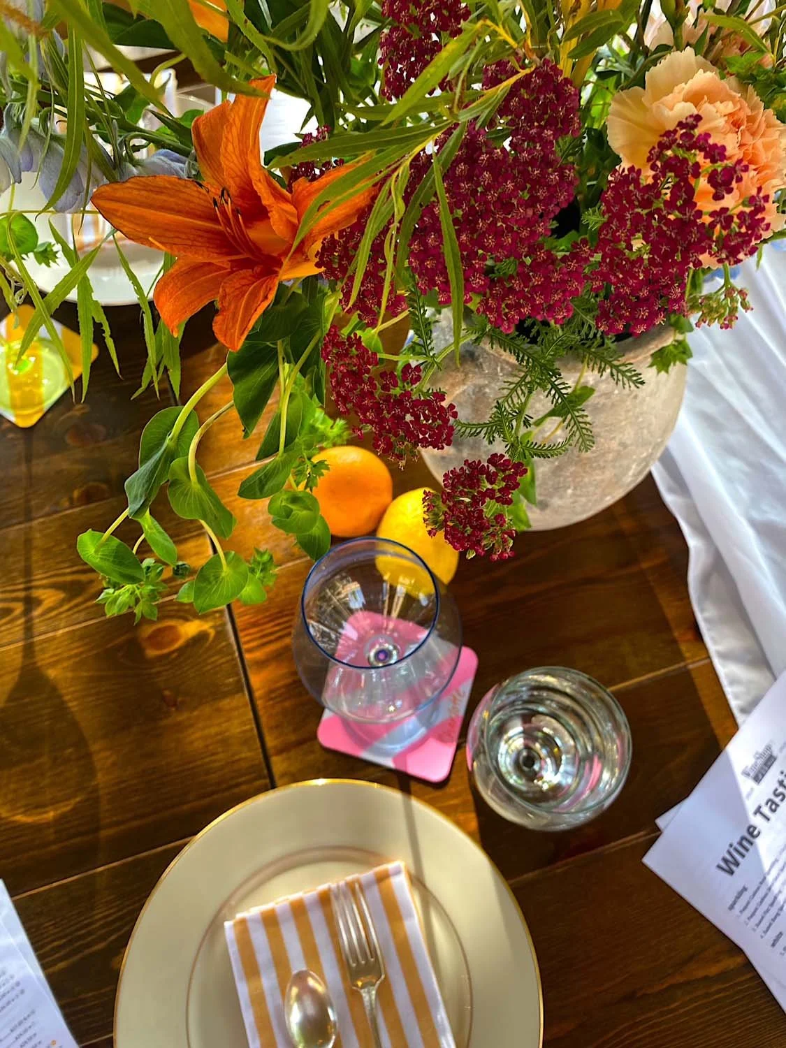 A wooden dining table set with a large floral centerpiece in a stone vase, featuring orange lilies, pink and red flowers, and green foliage. There are two glasses, a lemon, a peach, a fork and spoon on a stripe napkin on a plate, and some documents o