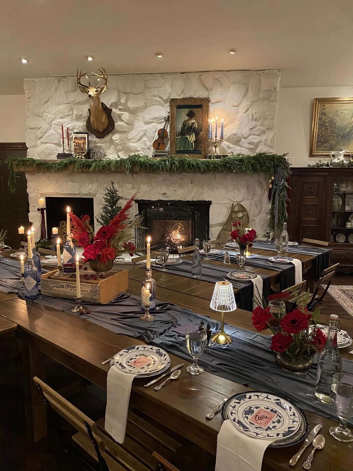 A decorated dining room with a long wooden table set for a festive meal, featuring red flowers, candles, and tableware. The background includes a stone fireplace with a green garland, a deer head mount, artwork, and a violin, with warm lighting.