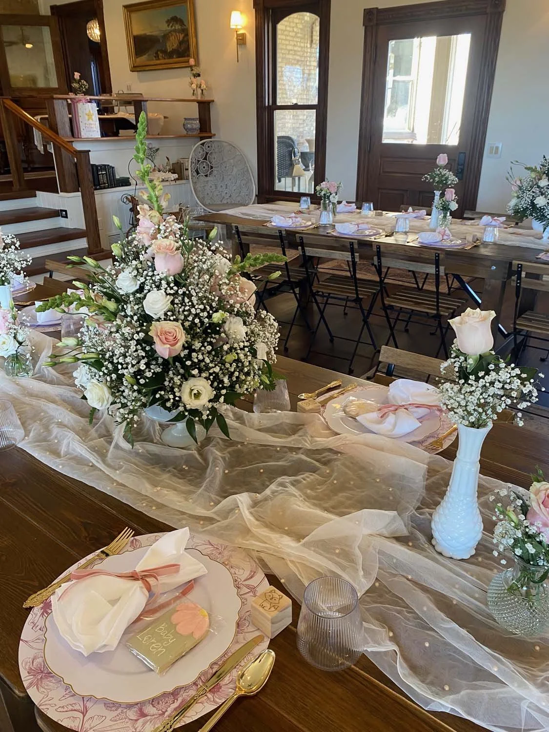 Decorated dining table with floral centerpieces, table settings, and pink accents in a cozy, well-lit room with wooden furniture and a view of an outdoor patio.