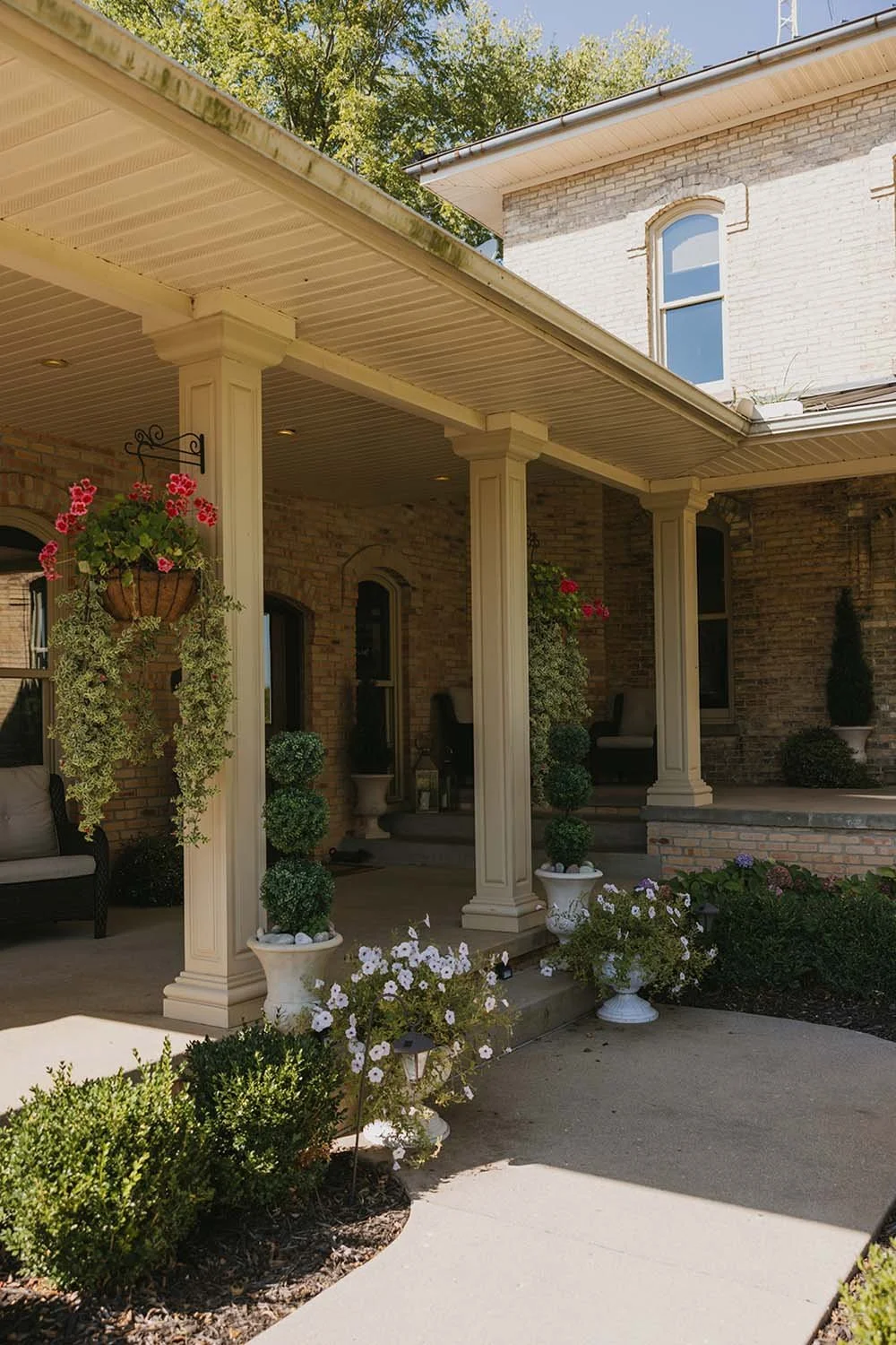 Front porch of a brick house with cream-colored columns, hanging flower baskets, potted plants, and garden flowers, with trees in the background.