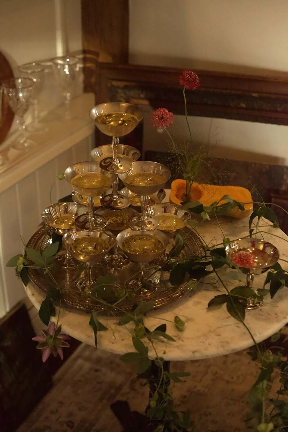 A pyramid of champagne glasses on a silver tray, decorated with green ivy and pink flowers, on a marble table. In the background, there is a wooden cabinet with empty glassware and a yellow cloth. A small pink flower is also on the table.