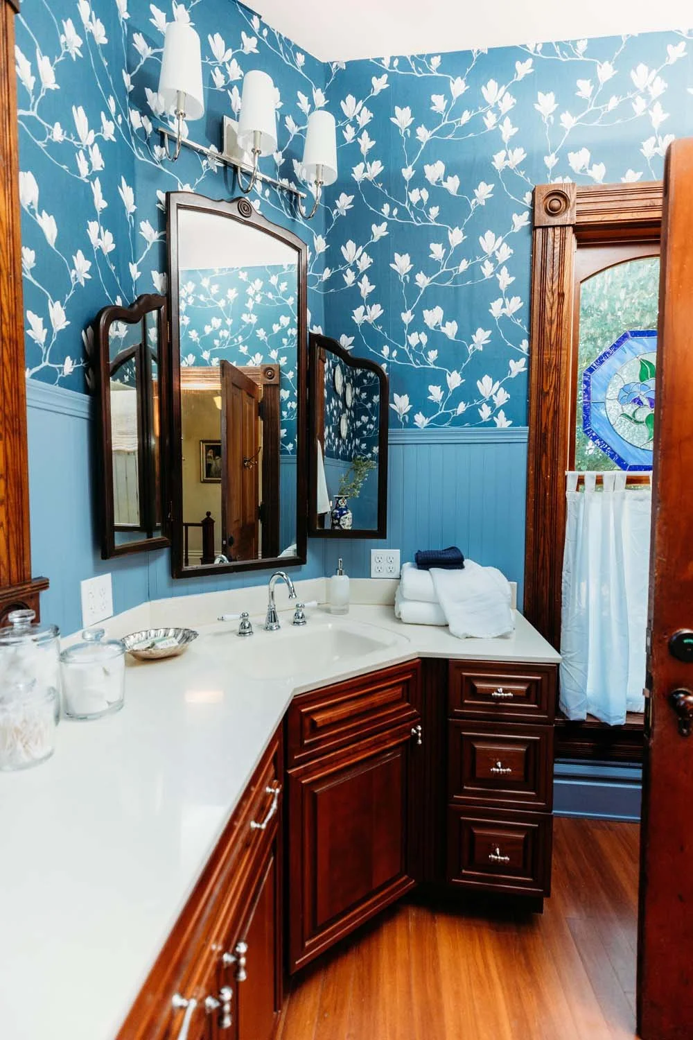 A vintage-style bathroom with a white countertop, dark wood cabinets, and triple mirrors above the sink. The walls are decorated with blue floral wallpaper and beadboard paneling. There's a stained glass window with a white curtain, and folded towels