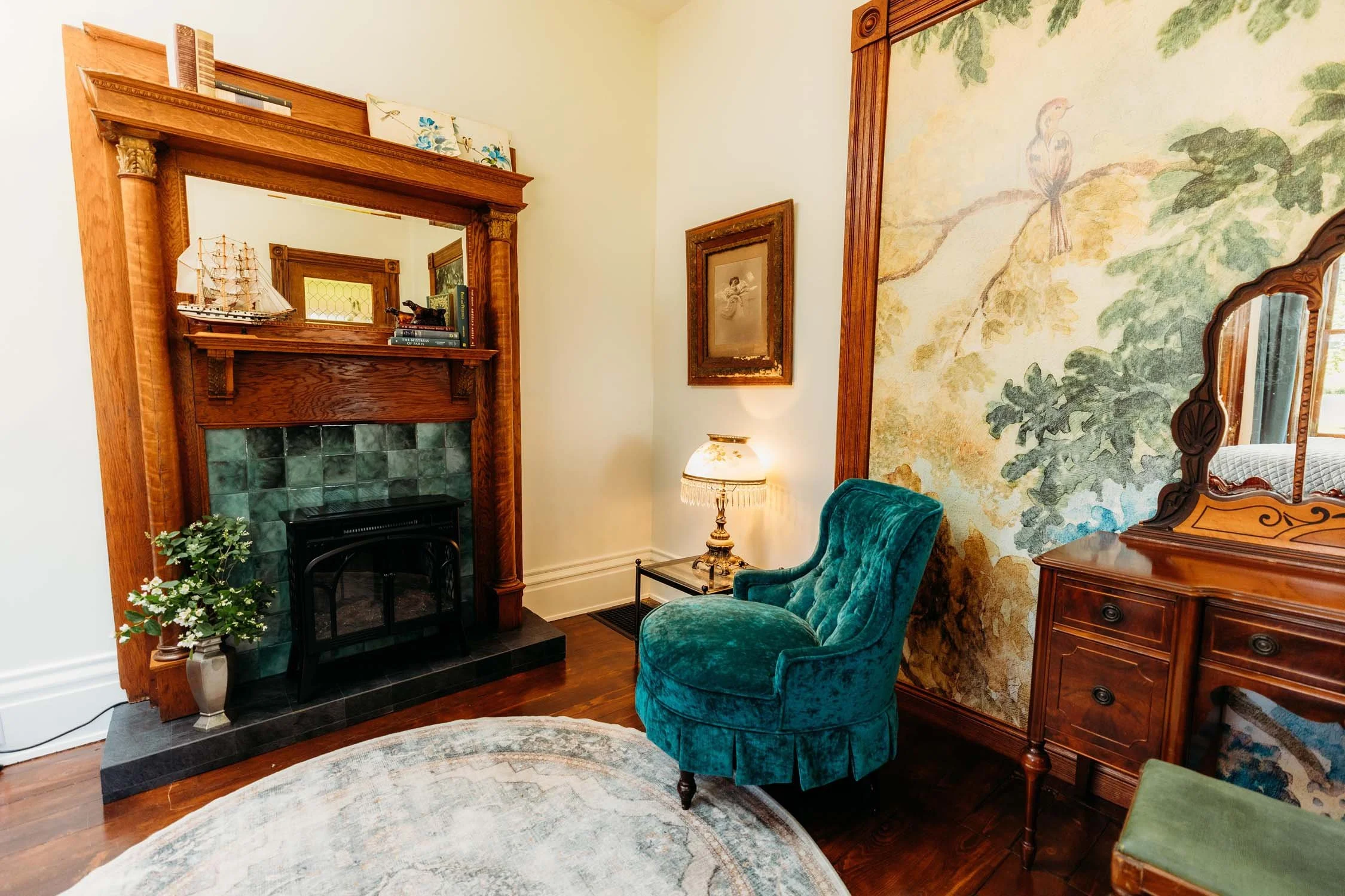 Vintage living room with a green velvet armchair, a wooden dresser with a mirror, a fireplace with decorative tiles, a painting, a table lamp, a framed picture, and a rug.