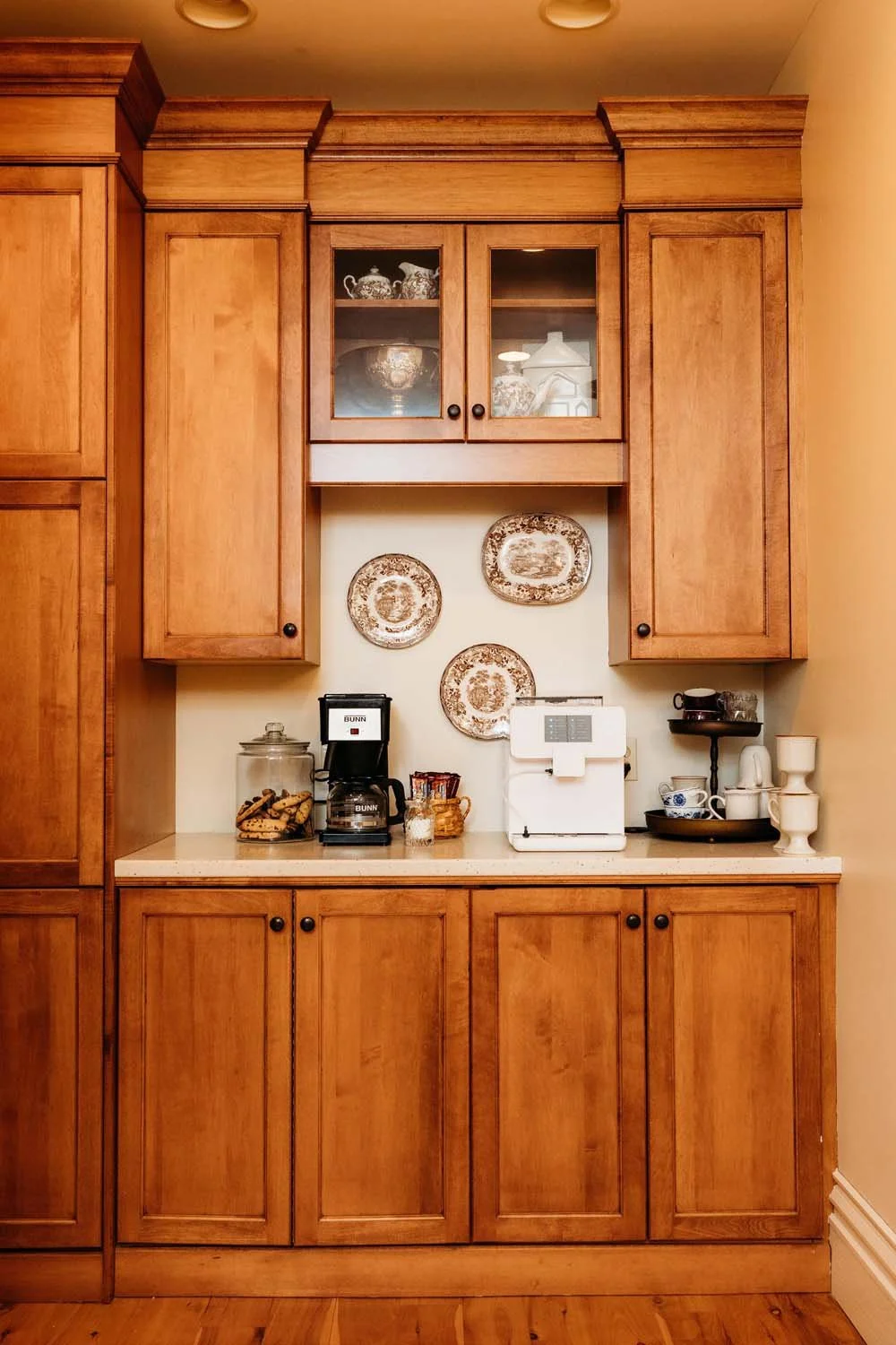 A kitchen cabinet with a coffee maker, a cookie jar, and some dishes on the countertop. Decorative plates hang on the wall behind. The cabinet is made of wood with black knob handles.