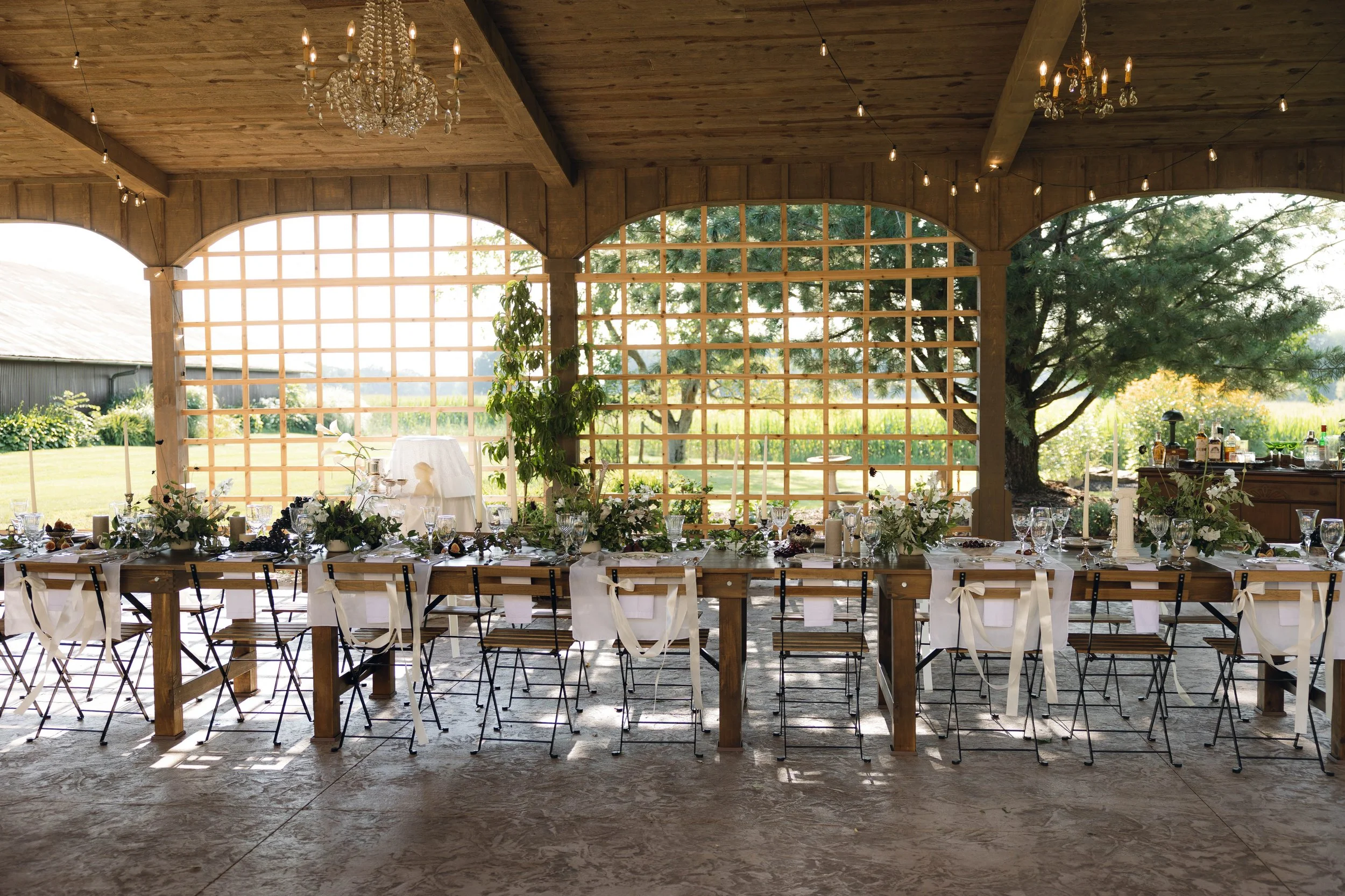Wedding reception table set up with floral arrangements, candles, and glassware in a rustic open pavilion with wooden beams and large windows overlooking a garden.