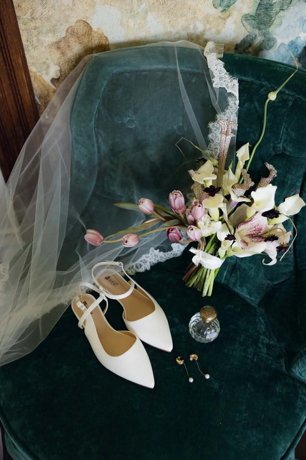 Wedding shoes, a bouquet of flowers, and perfume on a green velvet armchair.