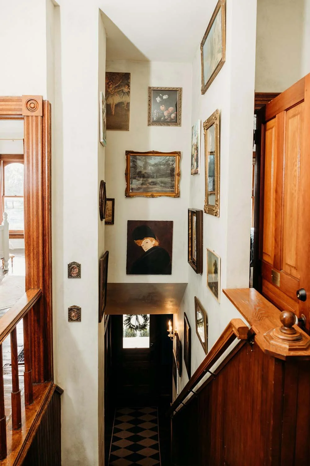 Interior view of a stairwell with framed artwork on the walls, a dark wooden staircase, and a chandelier at the bottom leading to a door with a window decorated with a wreath.