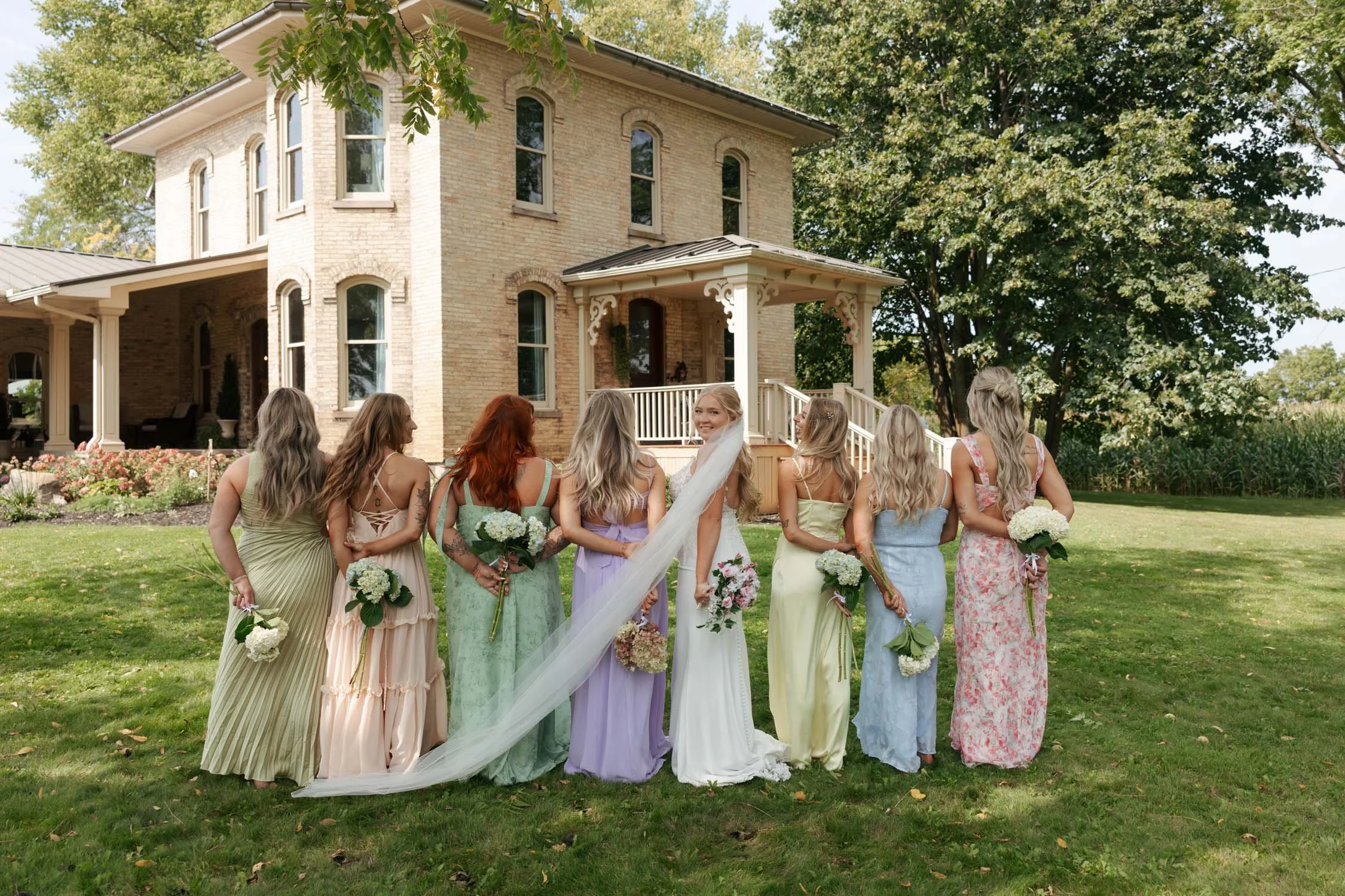 A bride and eight bridesmaids standing with their backs to the camera in front of a large brick house with a porch and trees. The bride is in a white wedding dress with a veil, and the bridesmaids are in pastel-colored dresses, each holding a bouquet