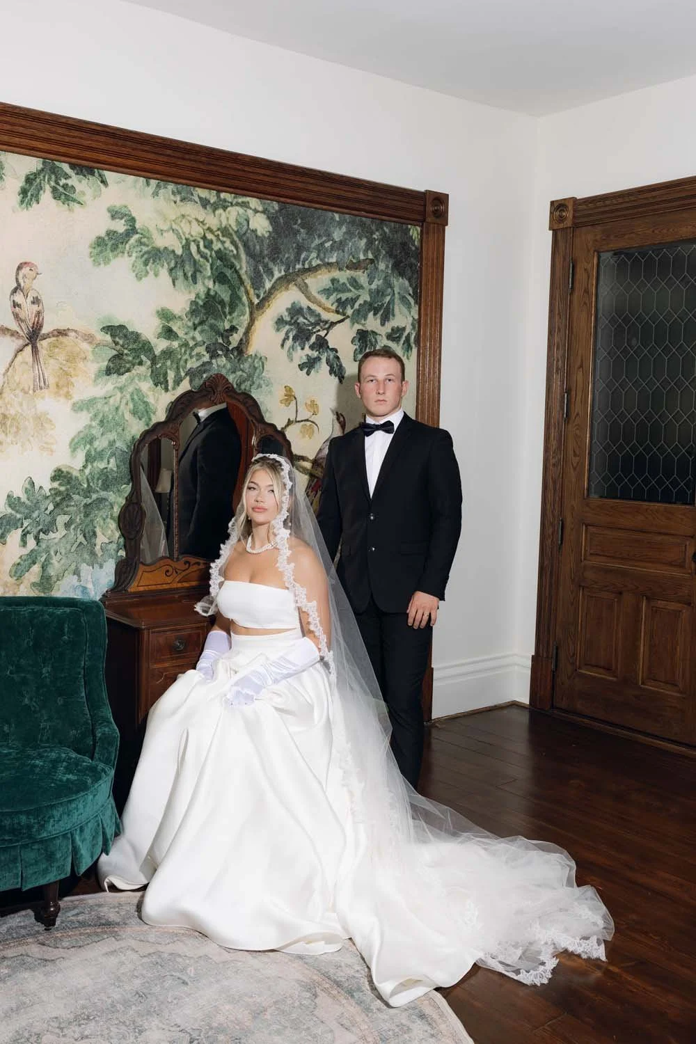 A bride in a white wedding dress with a veil and gloves, sitting on a green chair, and a groom in a black tuxedo standing beside her, inside a room with wooden accents and a mural on the wall.