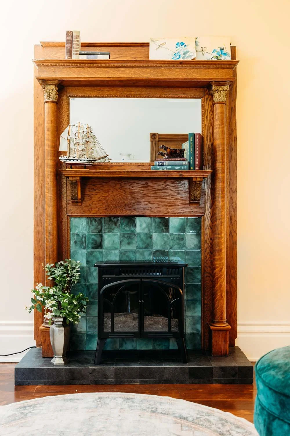 A fireplace with a wooden mantel and green tiled surround, decorated with a model sailboat, a stack of books, a small sculpture, and a decorative item with a floral pattern. There's a potted plant with white flowers on the left side, a mirror above t