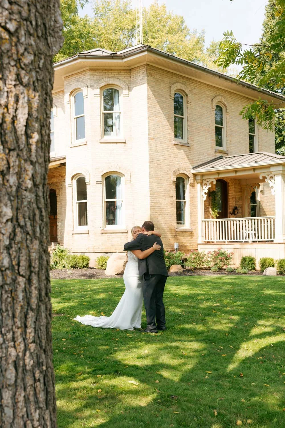 A bride and groom embracing in front of a large brick house with a porch and multiple windows, surrounded by green grass and trees.