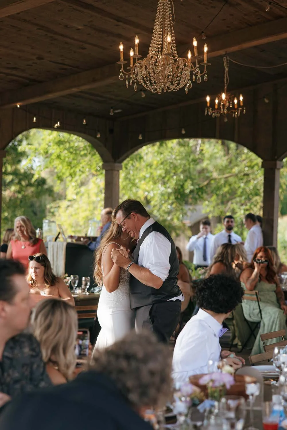 A bride and groom are dancing at their outdoor wedding reception under a wooden pavilion with chandeliers, surrounded by seated guests enjoying the celebration.
