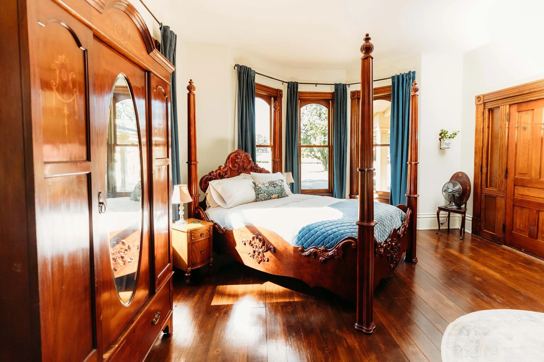 A bedroom with a four-poster wooden bed, dark blue curtains, and hardwood floors. There are windows with a view outside, a small wooden nightstand with a lamp, a vintage fan, and a large wooden wardrobe.