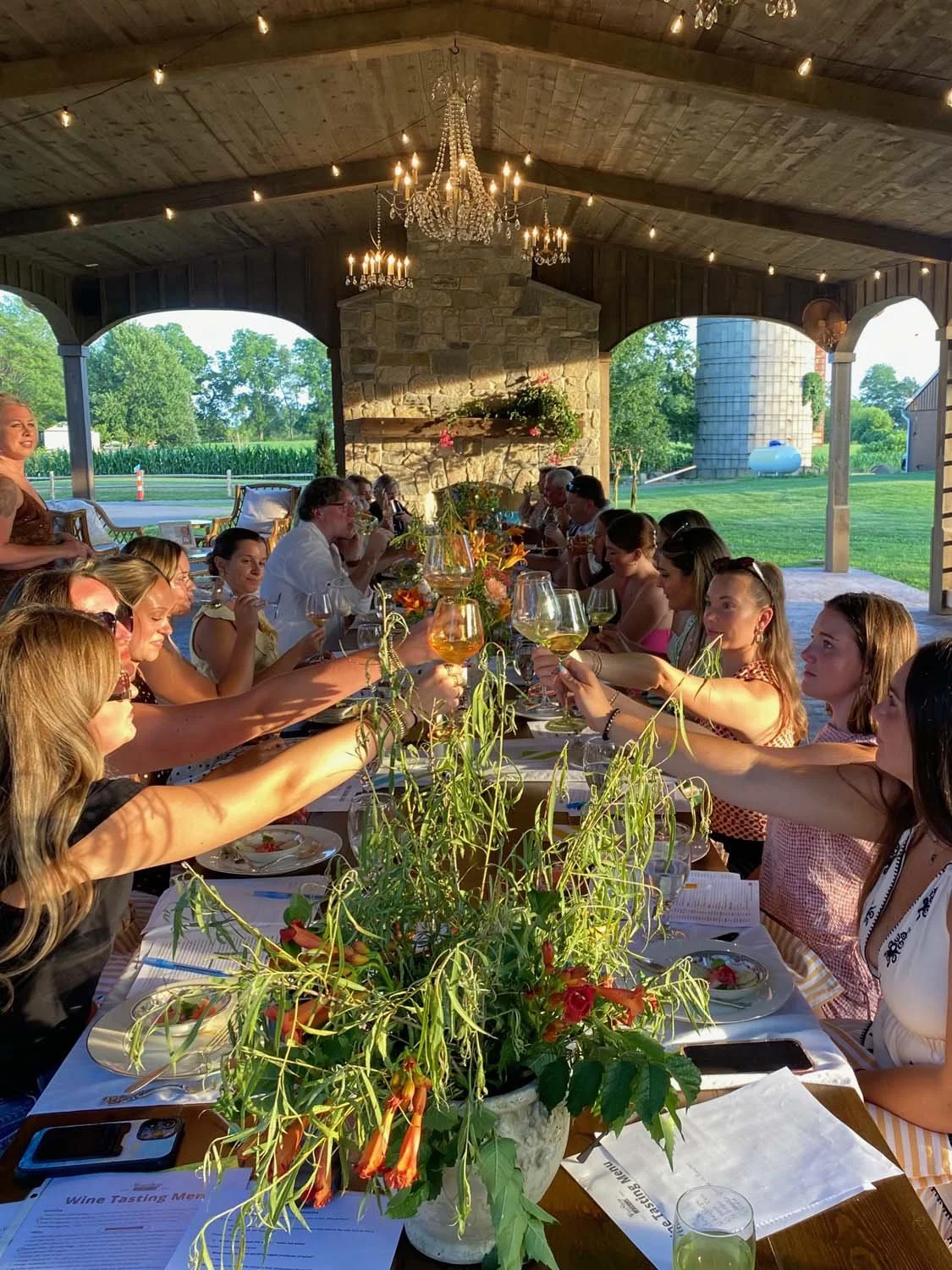 A group of people sitting around a long table outdoors, raising glasses in a toast during sunset, under a pavilion with chandeliers and string lights, with a rural setting in the background.