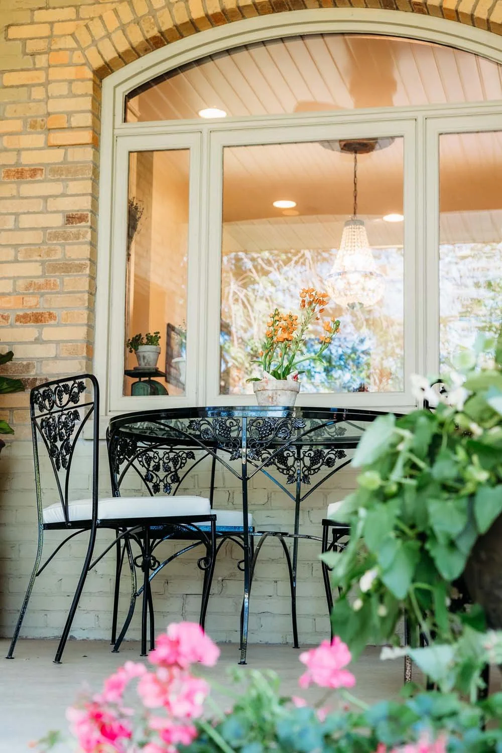 A cozy outdoor patio with black wrought iron table and chairs, decorated with potted plants and colorful flowers, viewed through a large window with a chandelier inside.