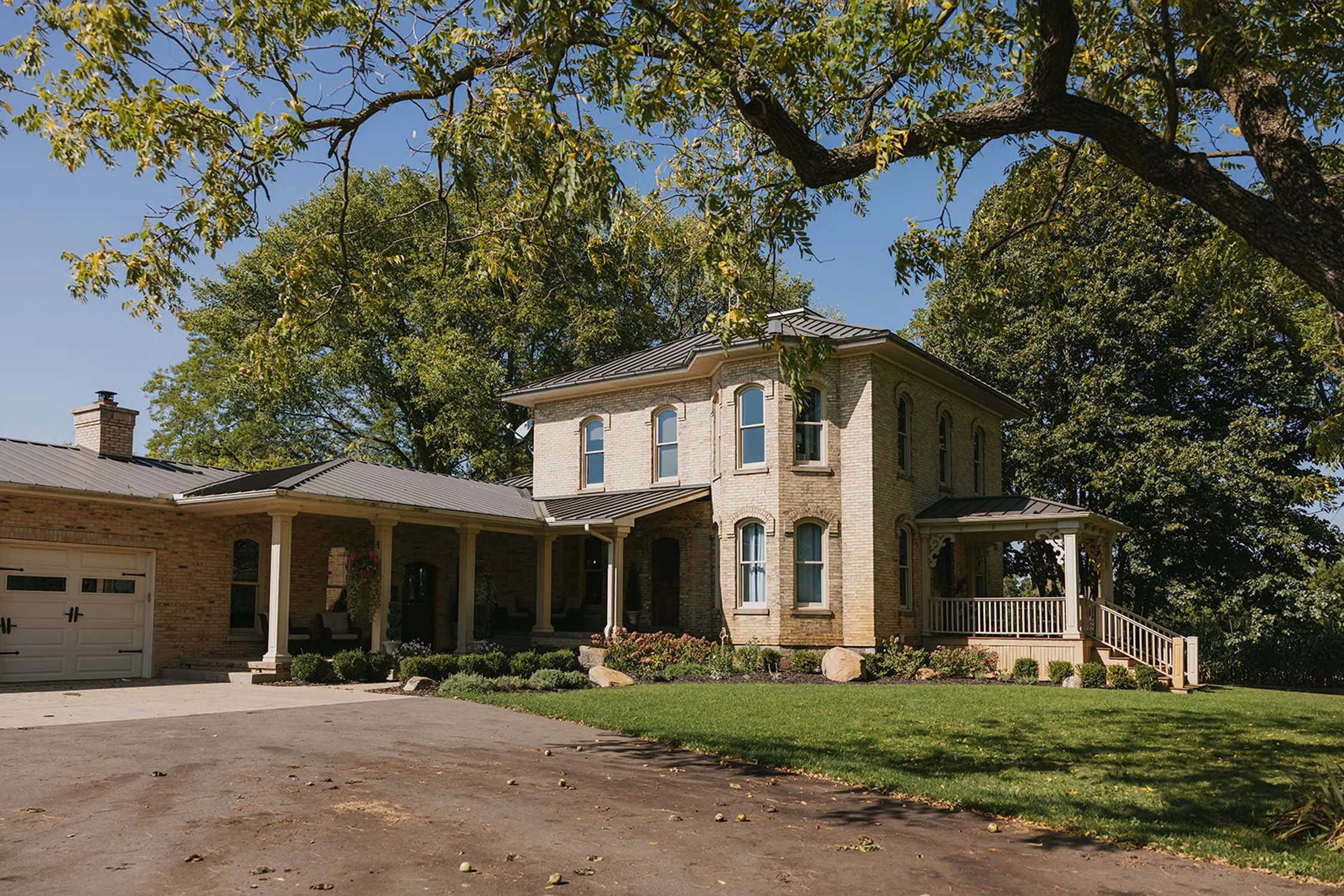 A two-story brick house with a metal roof, front porch, and landscaped yard surrounded by large trees.