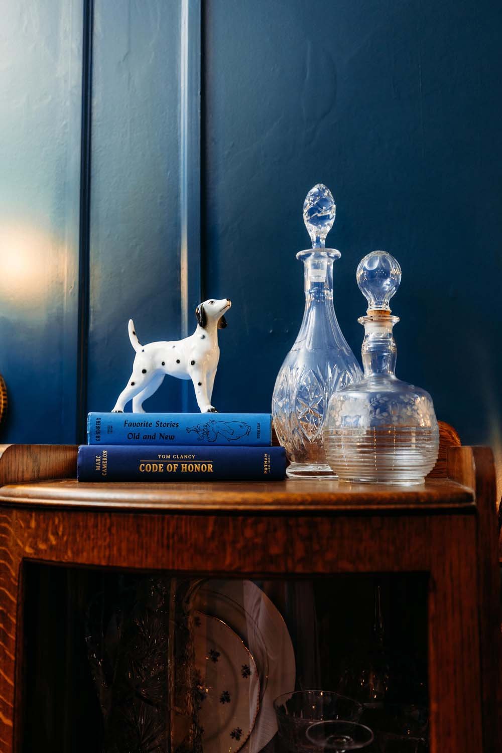 A wooden side table against a blue wall holds two glass decanters, a ceramic Dalmatian figurine, and two books, one titled "Favorite Stories Old and New" and the other "Code of Honor" by Tom Clancy.