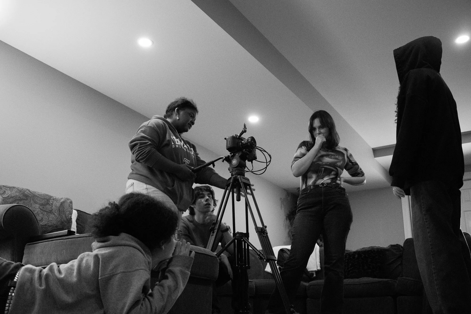 Group of young people on a film set with a camera and tripod in a living room. One person is operating the camera, another stands with a thoughtful pose, and three others are sitting or crouching, observing or preparing for a scene.