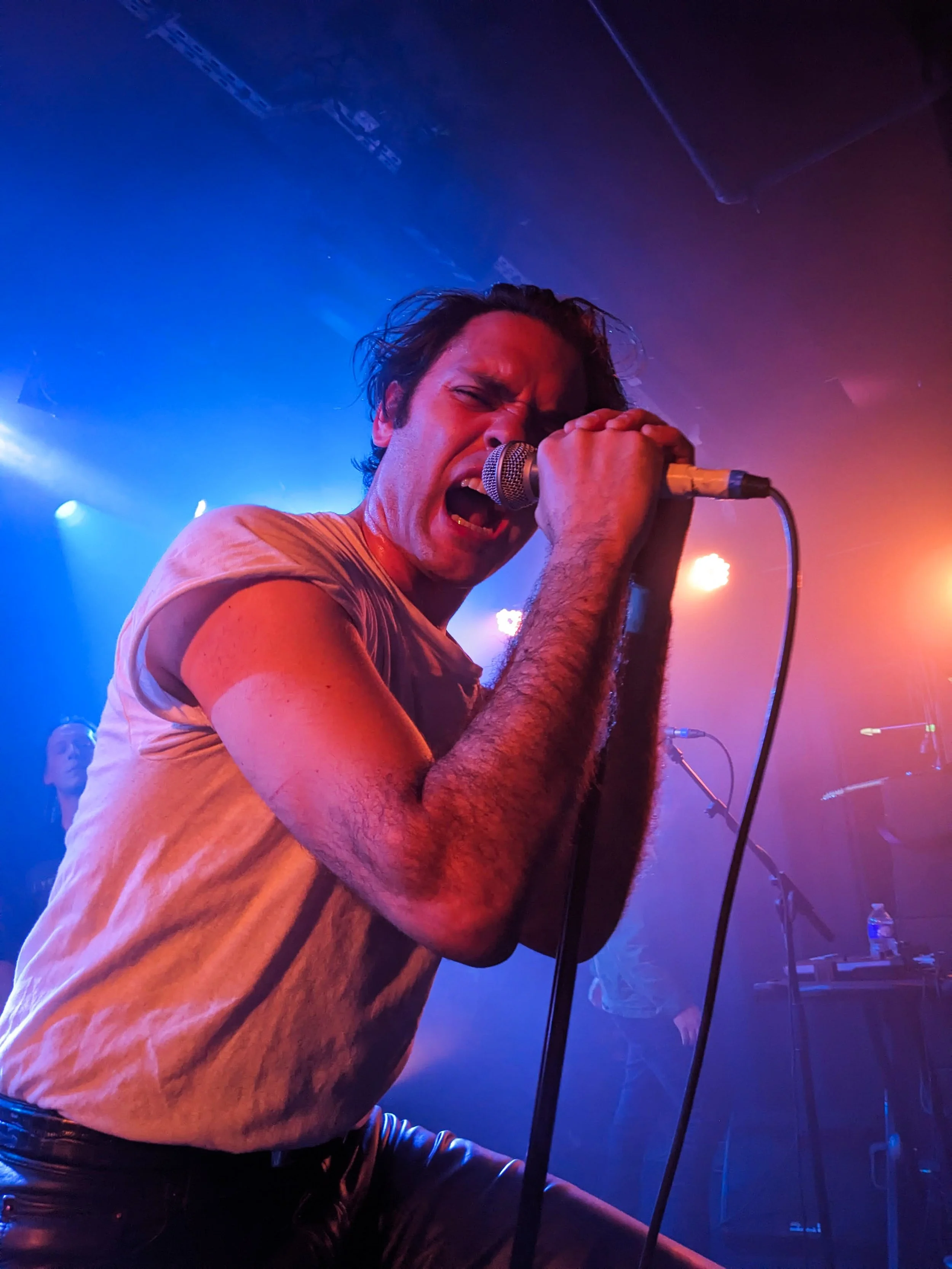 White male rock singer onstage, holding a mic and singing intensely. He is wearing a ratty white t-shirt and blue and red stage lights are shining through behind him.