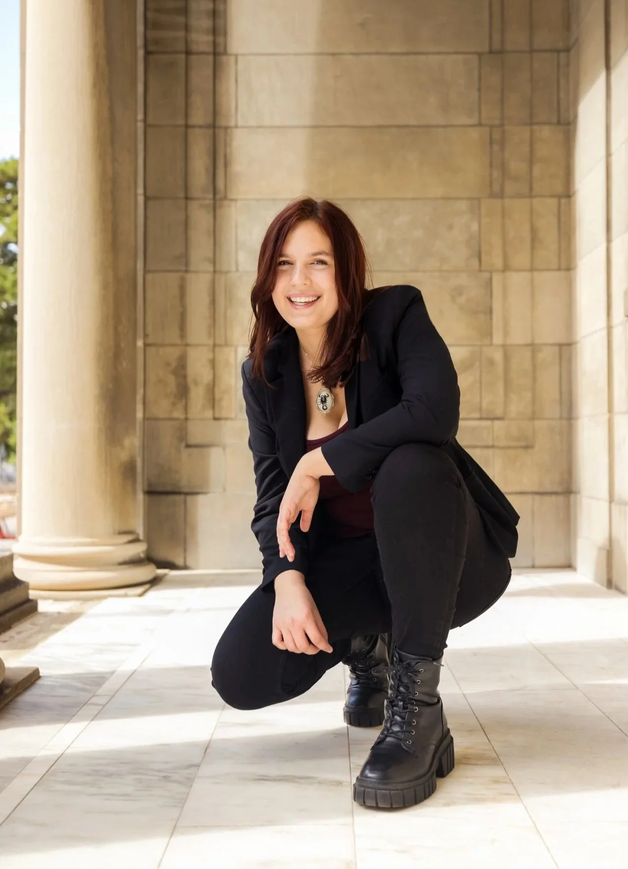 Casey Boyack, a twenty-something woman with medium-length auburn hair.  She is smiling, wearing a casual black suit and is crouched in front of a sandy stone wall in the sunlight.