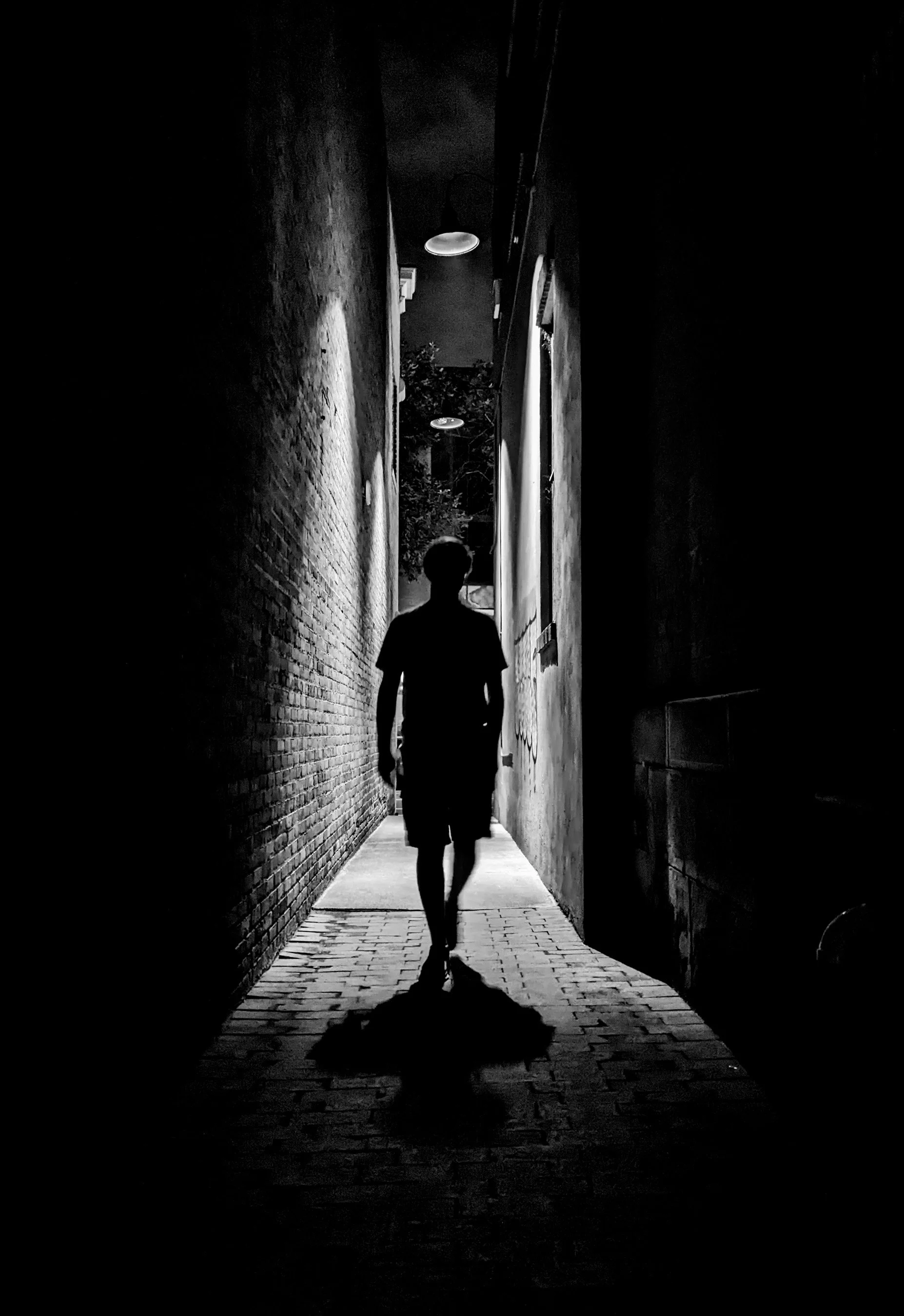 Black and white photo of a young man in shadow walking underneath an overhead light in a very dark and narrow alleyway