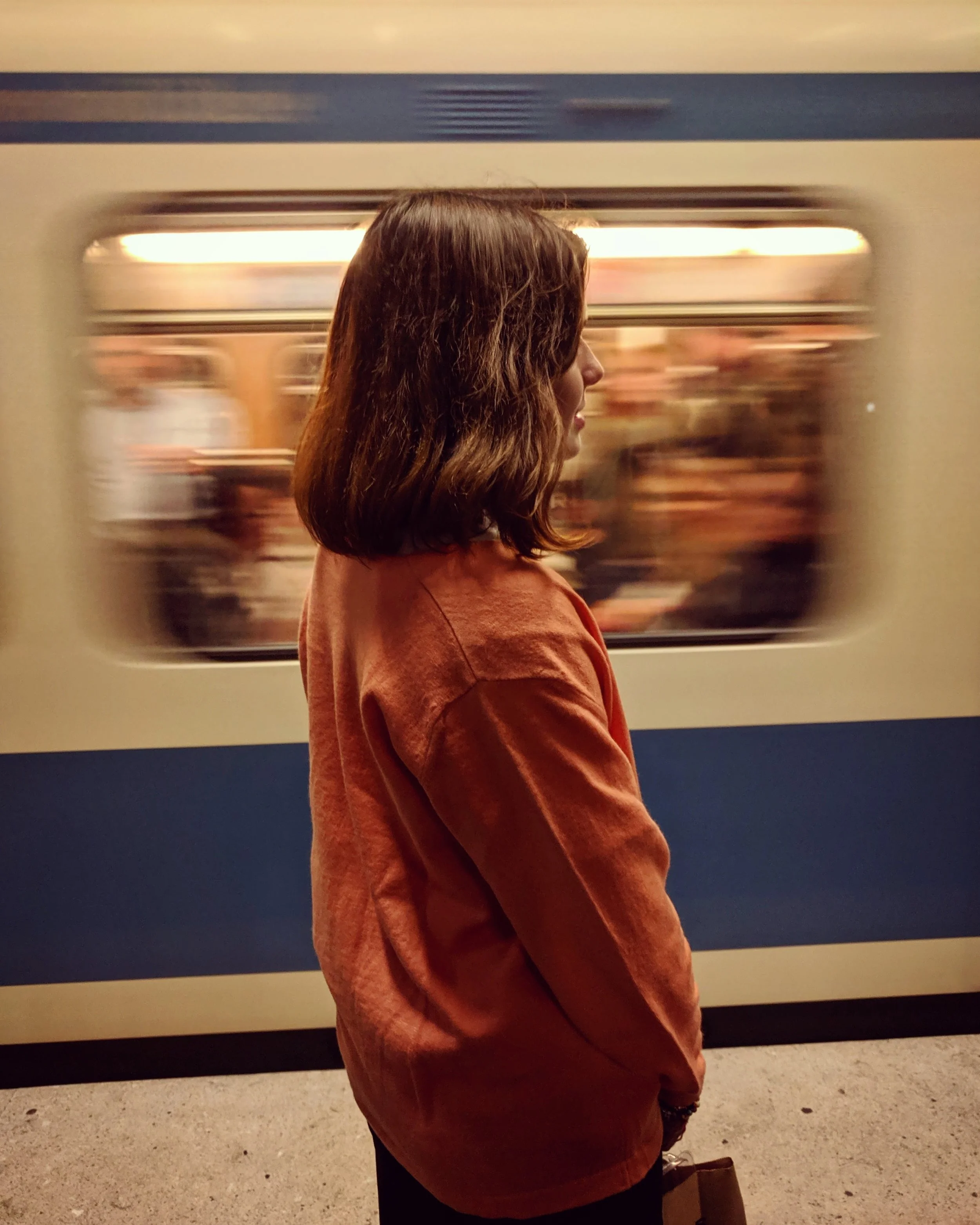 Profile of a white teenage girl with medium-length brown hair, wearing a rust-colored leather jacket. She is standing still while a white and blue striped subway train is flashing past her in the background. A blur of people can be seen through the t