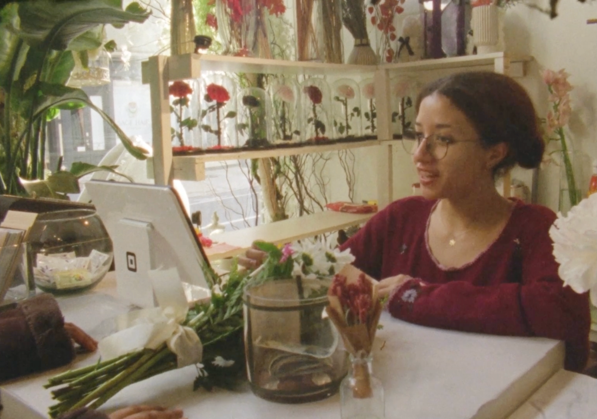 Young woman with tan skin, glasses, and dark hair that is pulled back in a bun, checking out a customer at a florist shop.