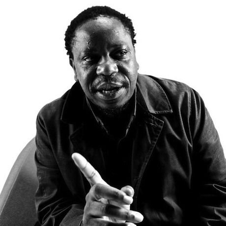 A black and white portrait of a man with short curly hair, wearing a dark jacket, looking at the camera, gesturing with his right index finger raised.