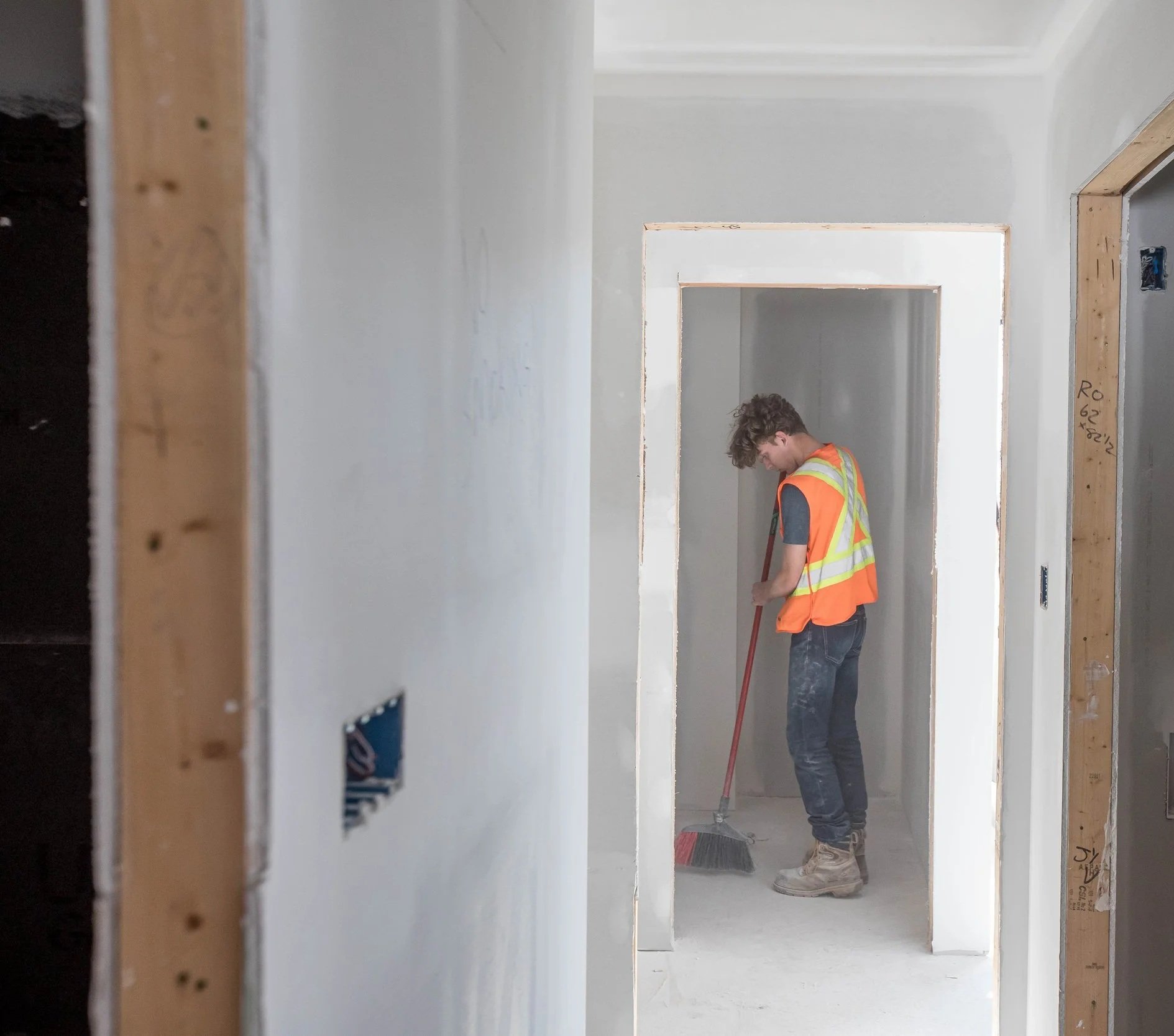 A young man wearing a construction safety vest sweeping a hallway in a house under construction.