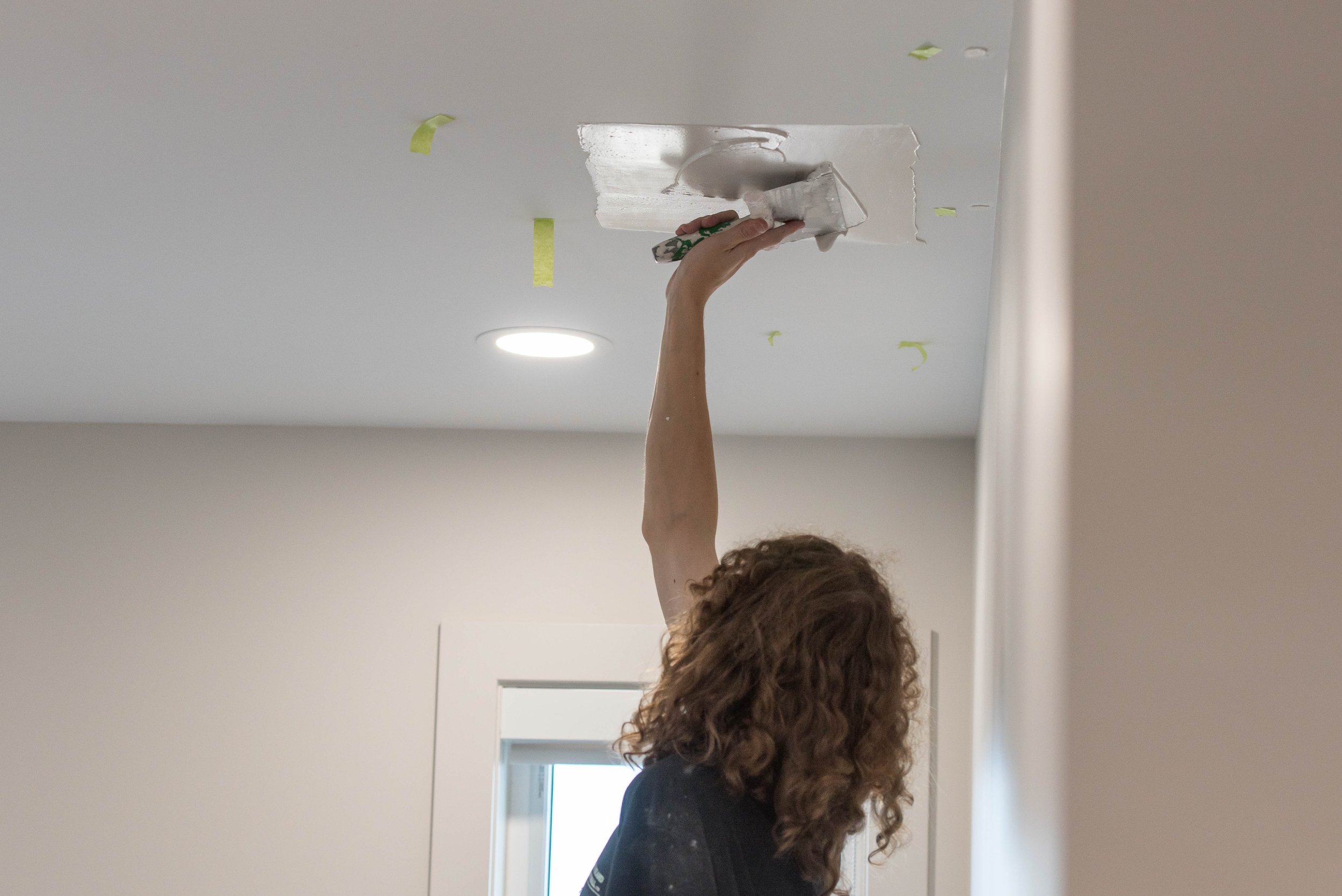 A person with curly hair painting a ceiling with a putty knife, with painter's tape on the ceiling around the area being painted.