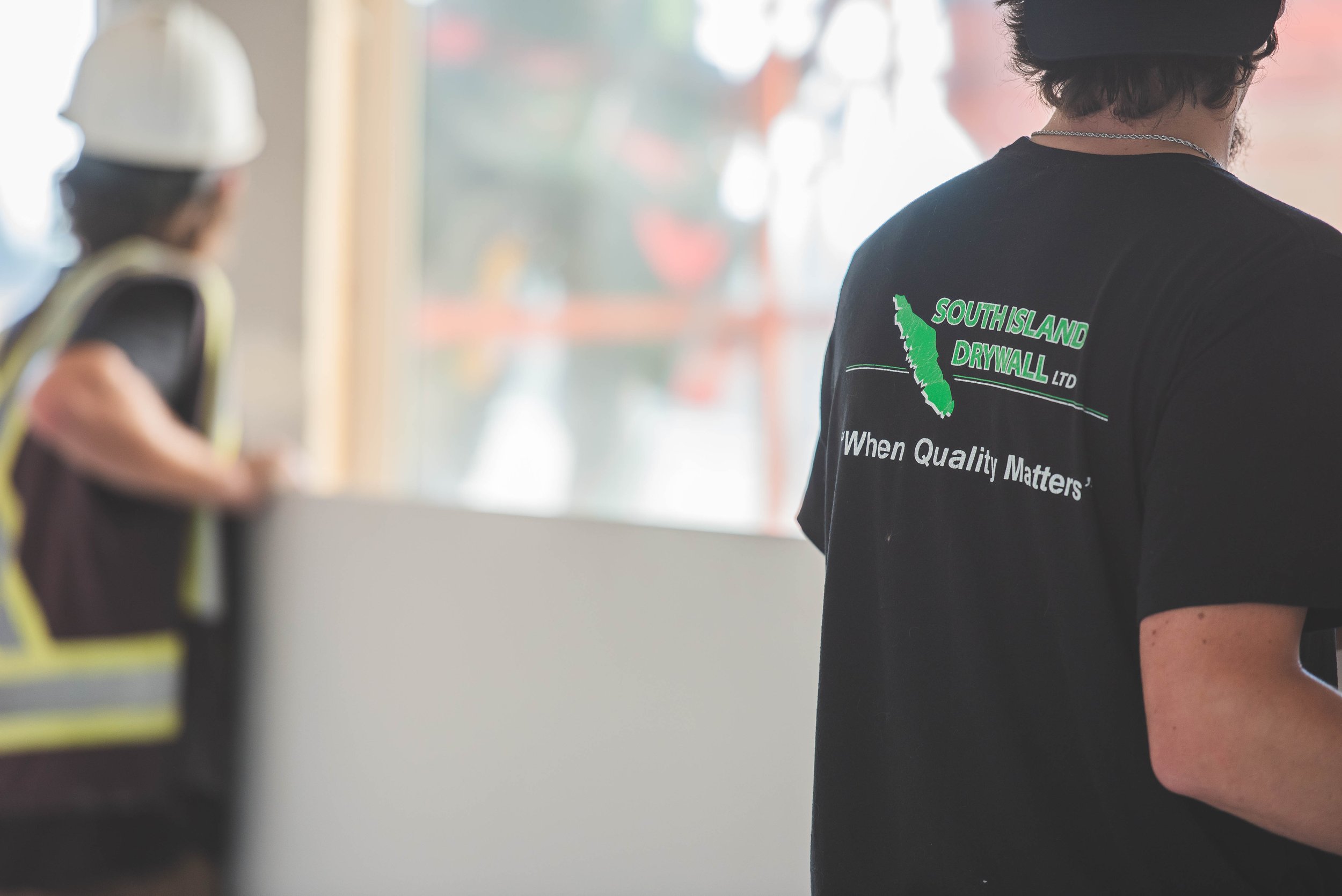 Person wearing a black t-shirt with green and white logo and text, standing inside a building near a large window with blurred construction worker in a yellow vest and hard hat outdoors in background.
