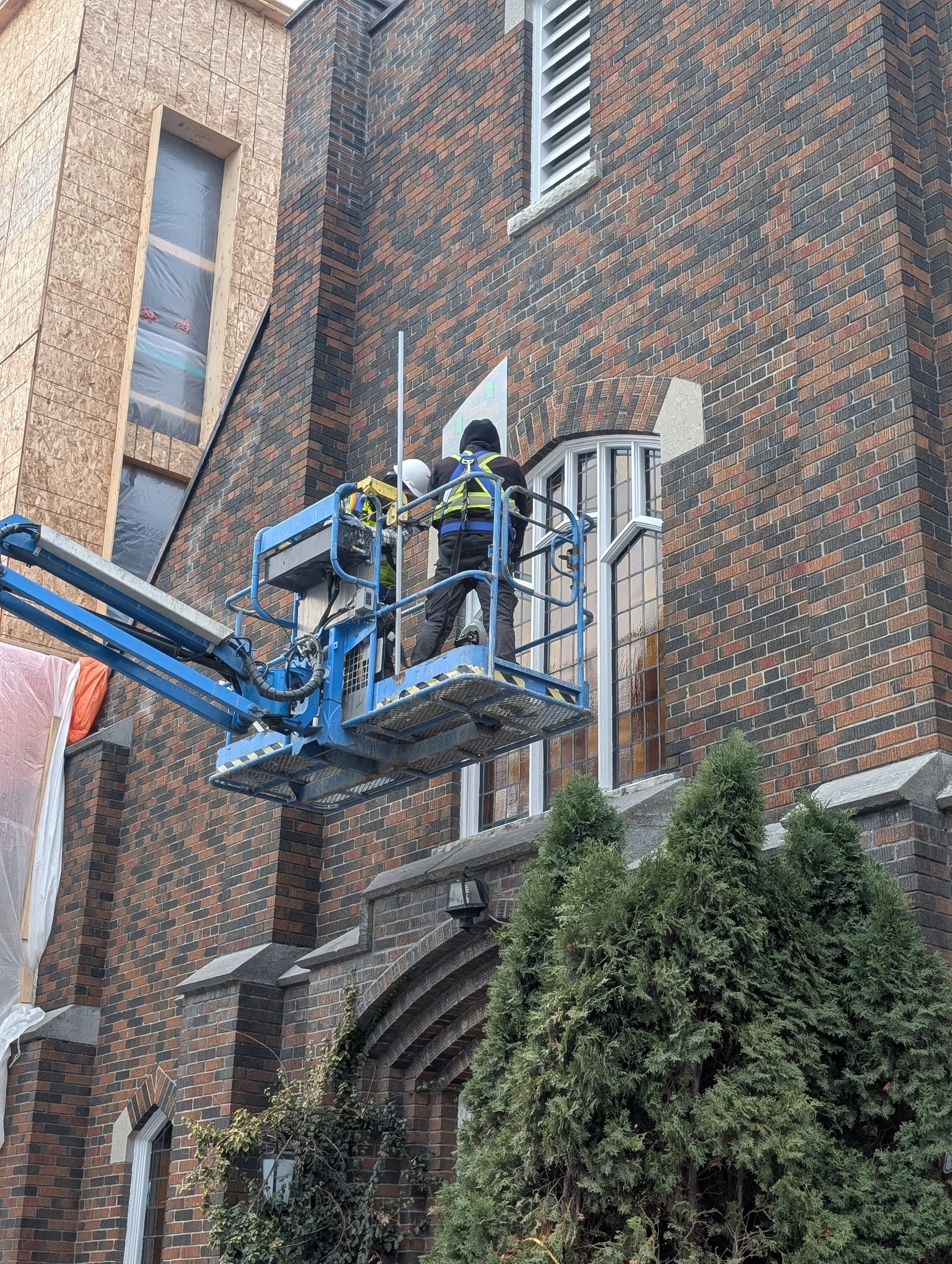 Two construction workers on a blue lift working on the exterior of a building with brick walls and a window.