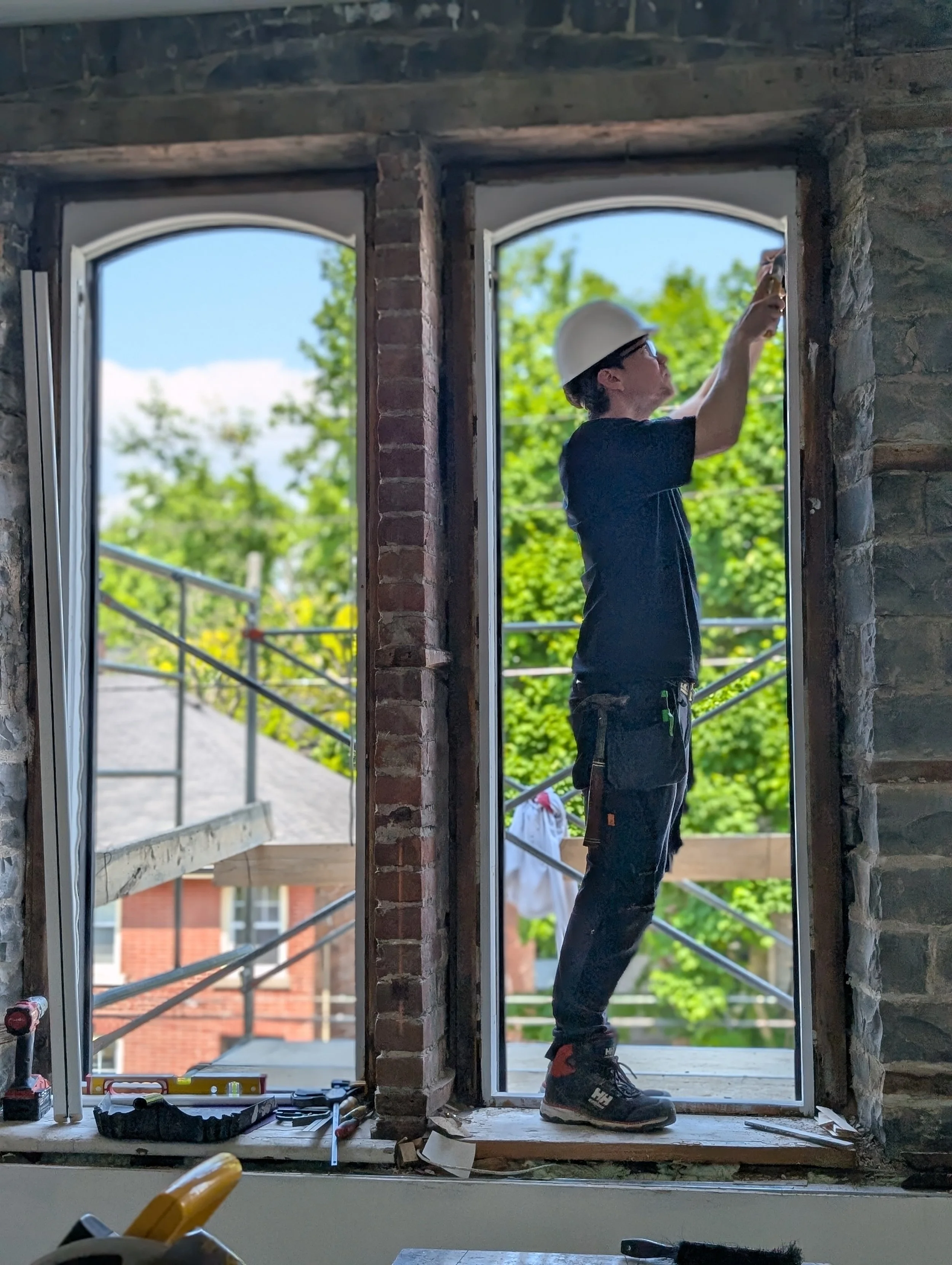 A construction worker wearing a white safety helmet and black work clothes standing inside a building, installing a large window with a view of green trees outside.