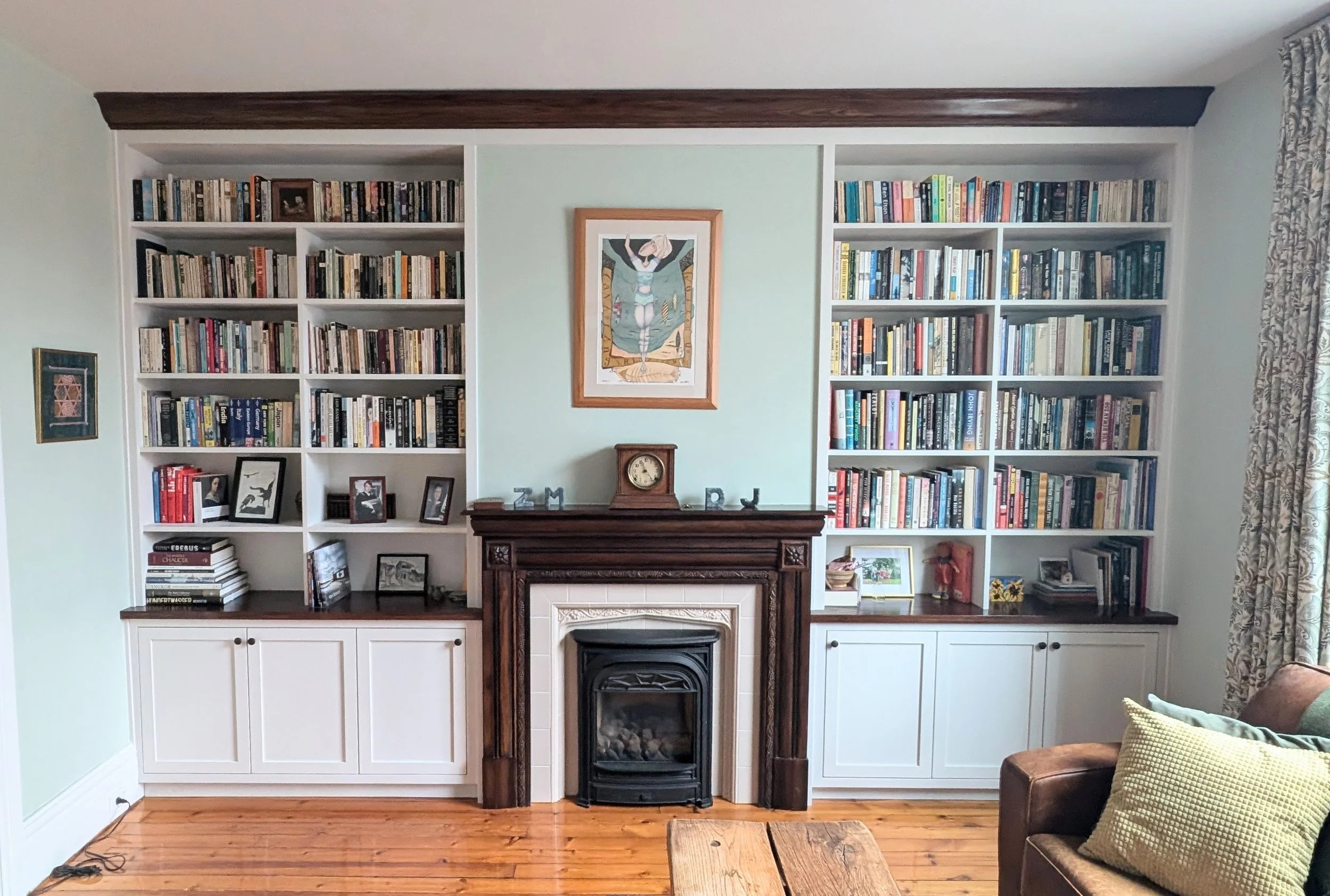 Living room with built-in white bookshelves, a wooden mantel fireplace, hardwood floor, framed artwork, set of decorative lights with letters 'Z', 'M', 'R', 'J' on the mantel, picture frames, books, and a brown leather sofa with pillows on the right.