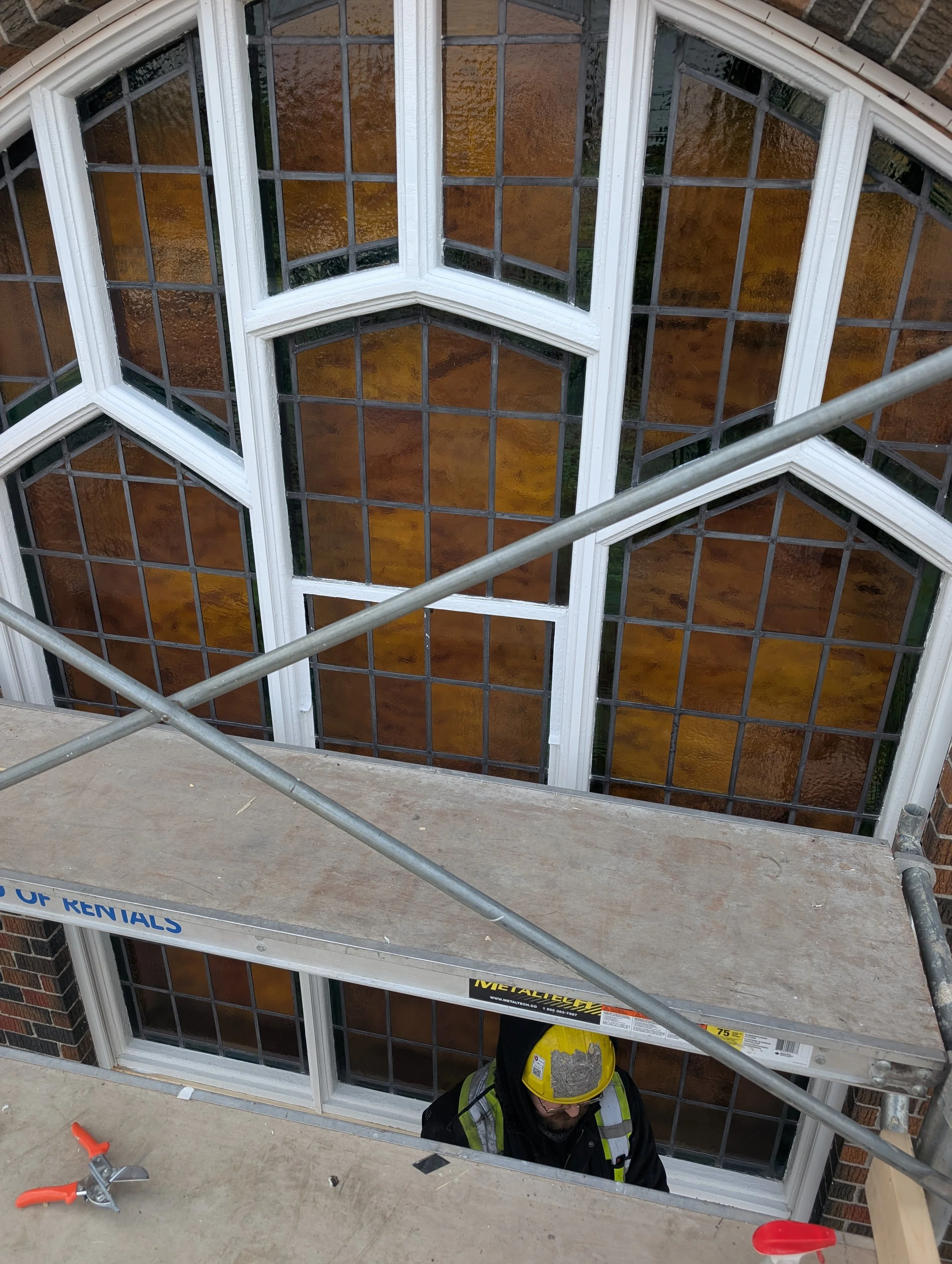 A construction worker wearing a yellow safety helmet and vest stands below a large, multi-paned stained glass window in a building under construction, with scaffolding and a work platform in the foreground.