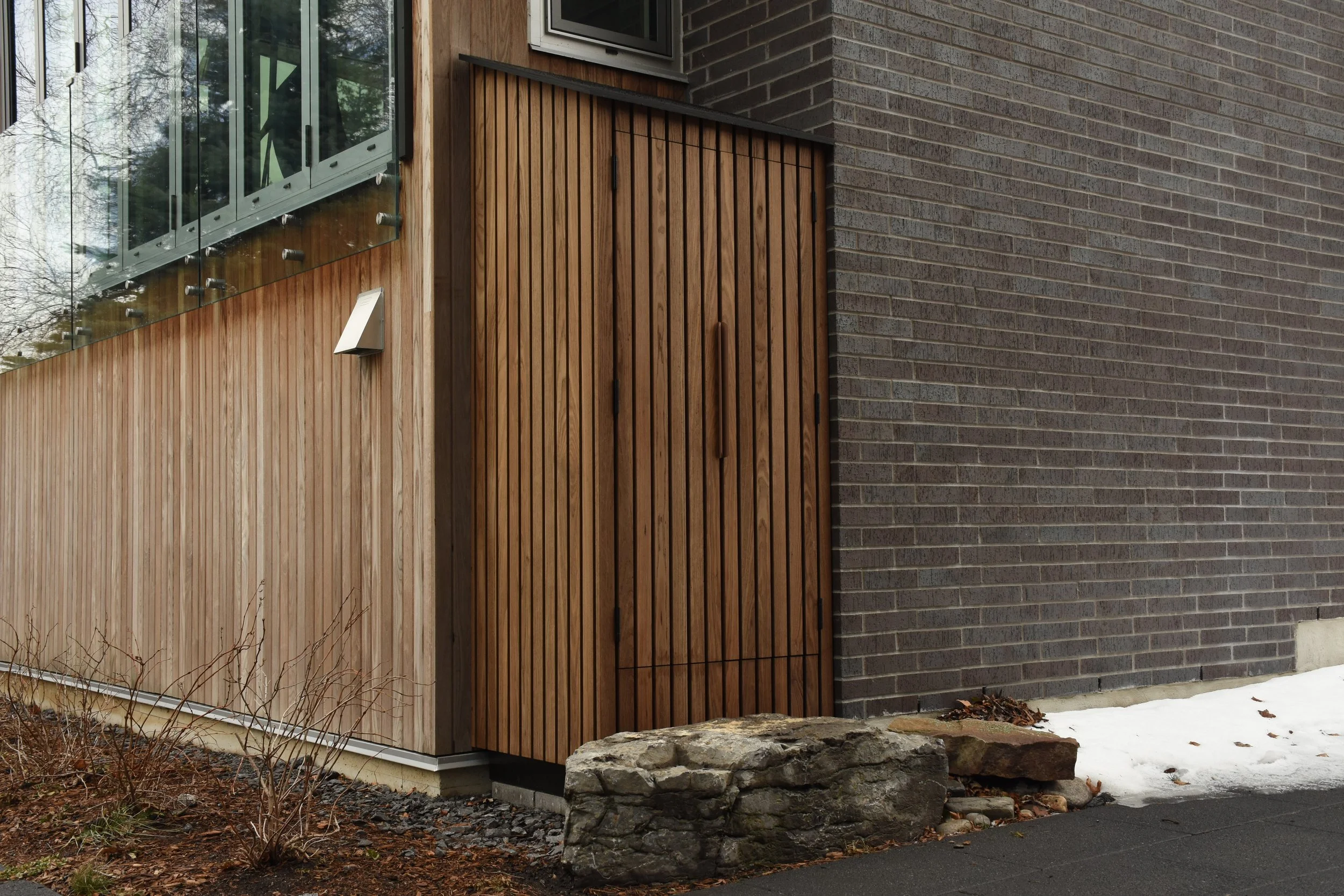 Exterior corner of a modern building with a mix of wooden and brick siding, a wooden door, and a small patch of snow on the ground.