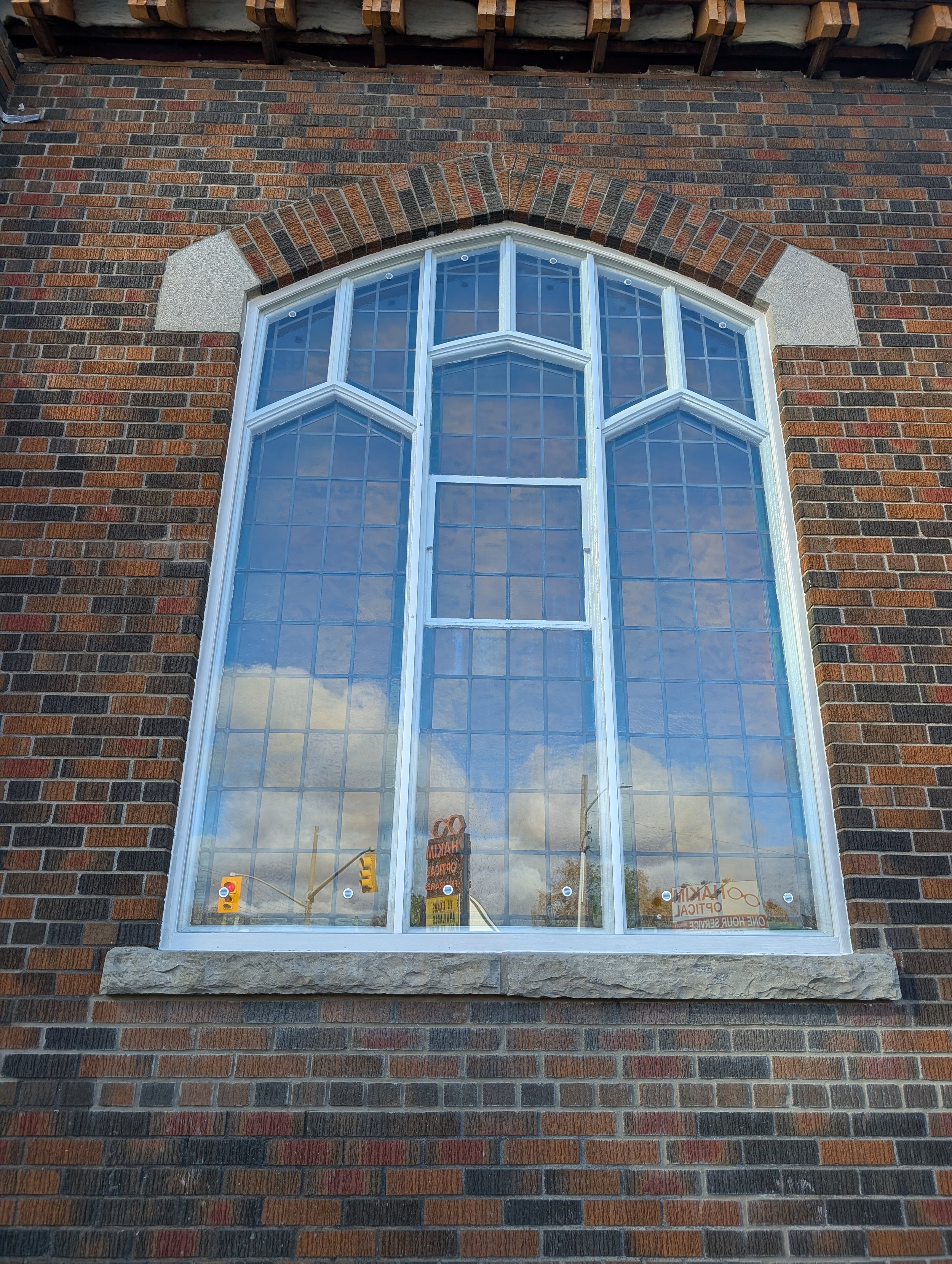 A large, decorative window set in a red and dark brown brick wall with a stone window sill. The window has multiple panes divided by white frames, with an arched top. The reflection of a street scene and partly cloudy sky can be seen in the glass.