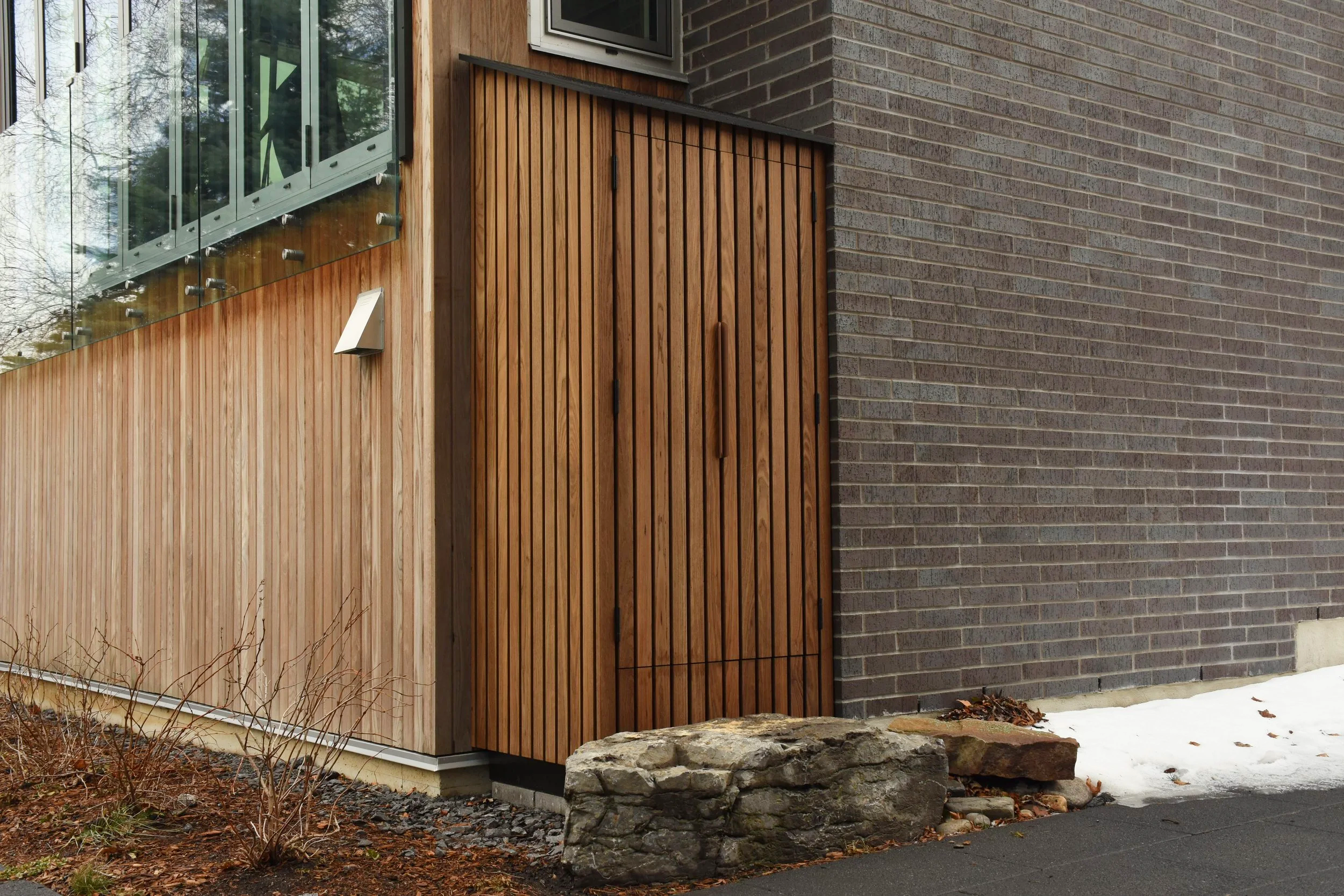 Close-up of a modern building corner with wooden vertical slat door, brick wall, glass window, and an external light fixture, with a rock and some snow on the ground.