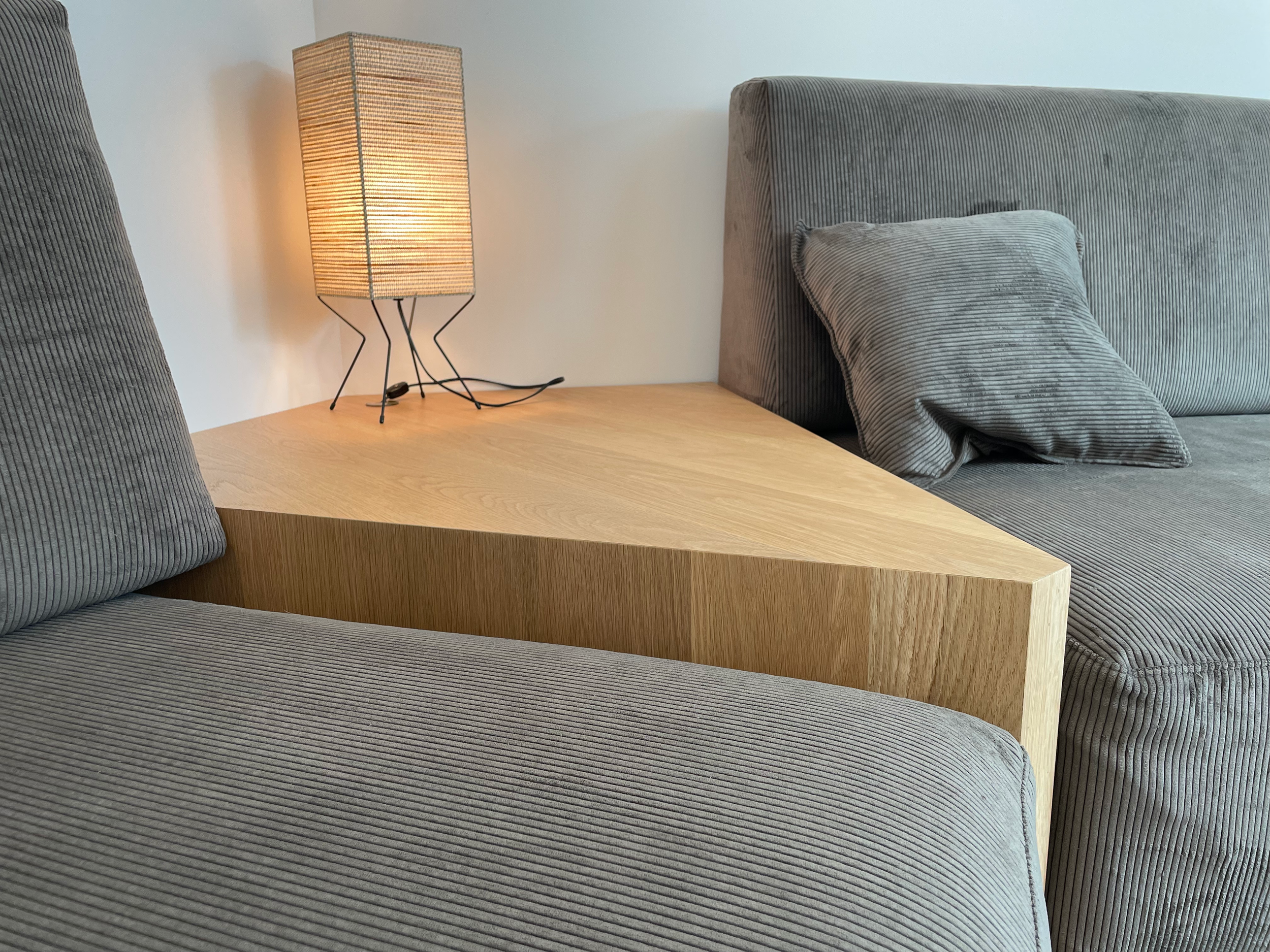 A modern living room corner with a beige triangular wooden side table, a gray corduroy sofa with matching pillows, a tall textured beige lamp on the table, against a plain white wall.